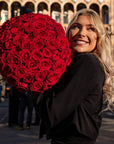 Luxurious dome-shaped bouquet of deep red roses held by blonde woman in elegant black coat, showcasing premium Valentine's Day arrangement against historic London architecture - Amaranté London