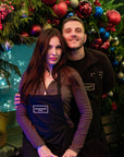 Two people wearing Amarante London aprons smiling in front of a festive Christmas wreath display during a luxury wreath-making workshop in London.