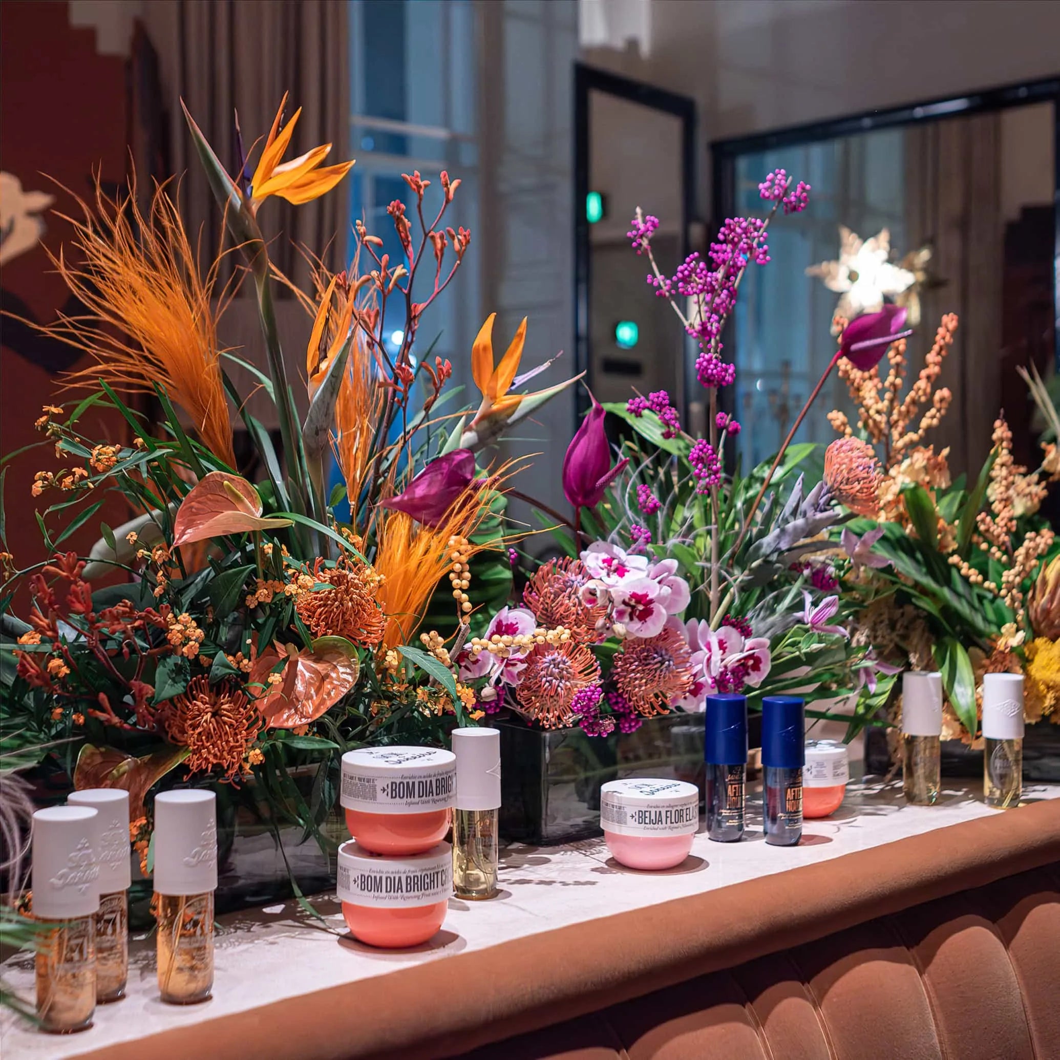 A lavish array of Sol de Janeiro's beauty products elegantly presented on a grand table amid Amaranté London's striking floral arrangements, with a backdrop of chic interior decor at Maine Mayfair restaurant.