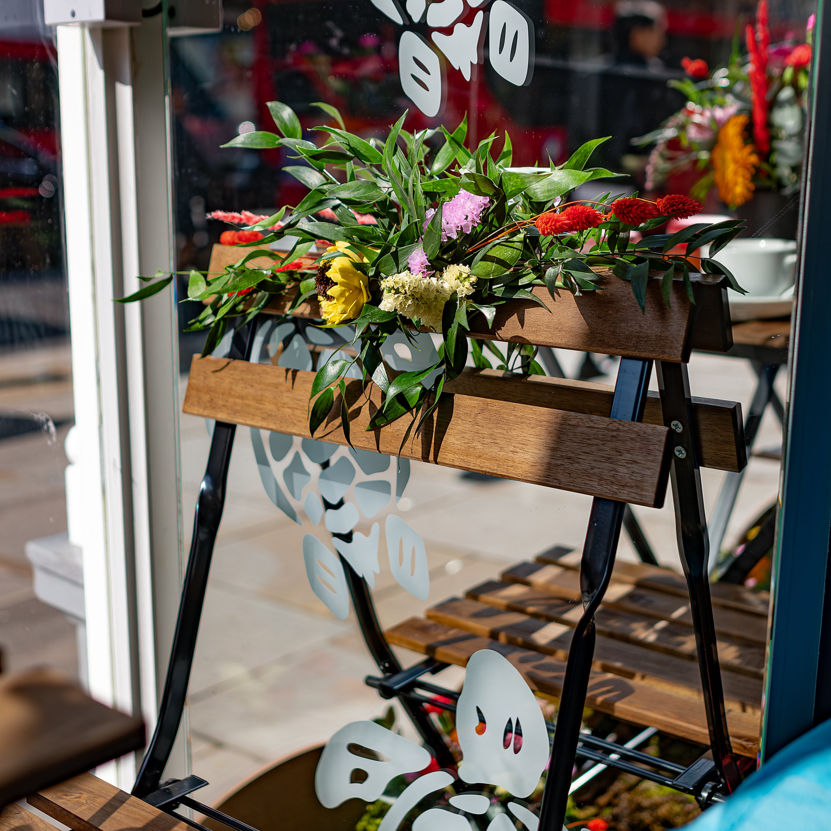 This is a close-up of a floral bench arrangement by Event Florist Amaranté London for The Vilebrequin Chelsea in Bloom display. It showcases a mix of red, yellow, and pink flowers with lush green foliage.