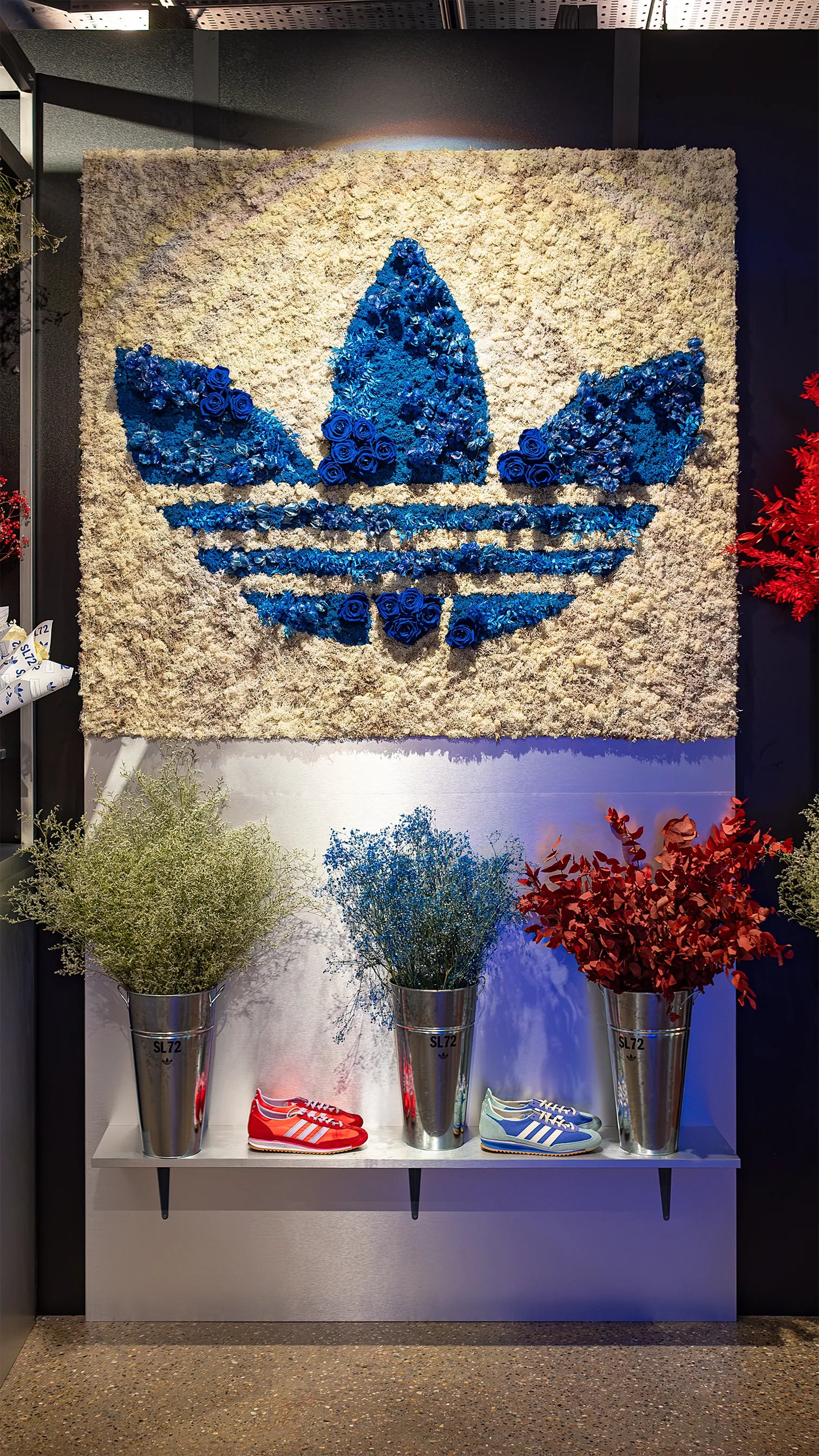 Bespoke floral brand logo installation featuring blue roses forming Adidas trefoil on textured floral backdrop with complementary preserved foliage arrangements - Amaranté London