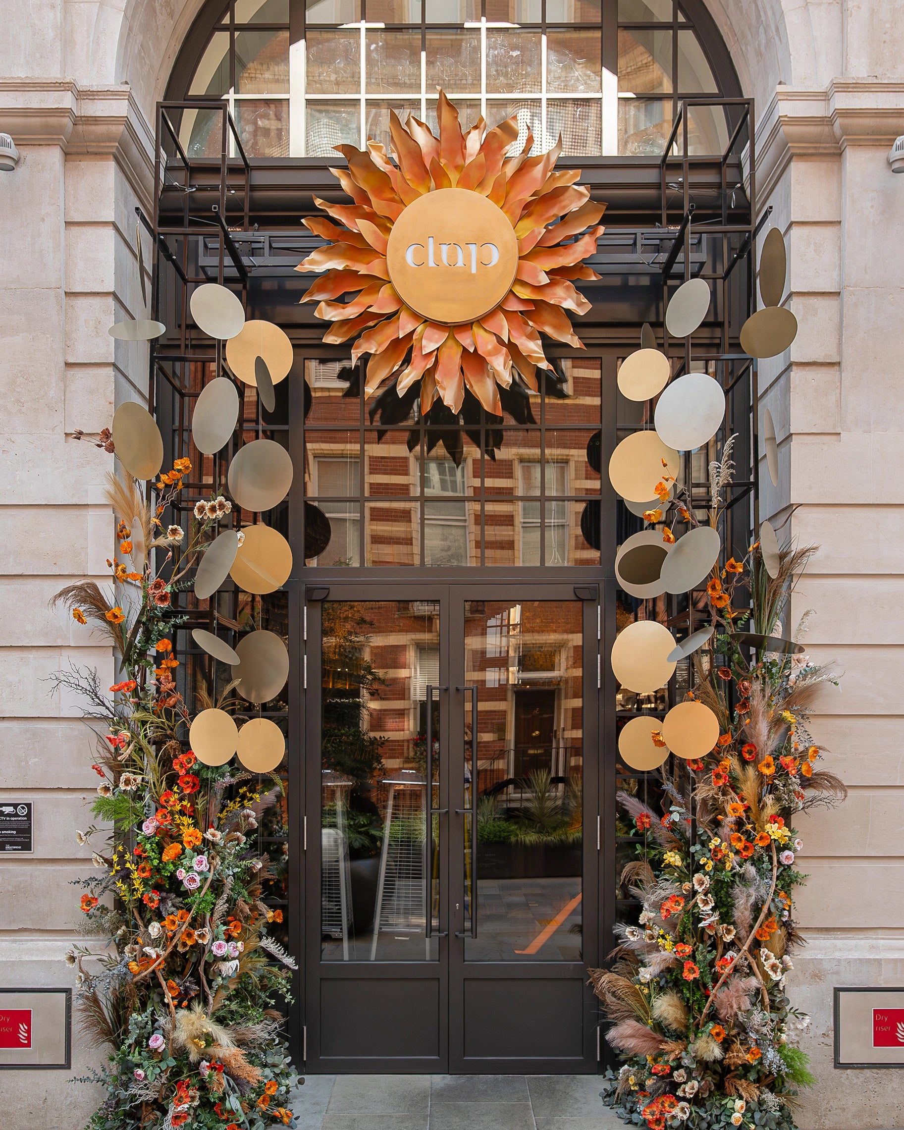 Seasonal floral installation featuring autumn blooms, pampas grass, and dried elements flanking venue entrance with sculptural sunburst design, showcasing Amaranté London's Winter Solstice arrangements at CLAP London - floral design by Amaranté London
