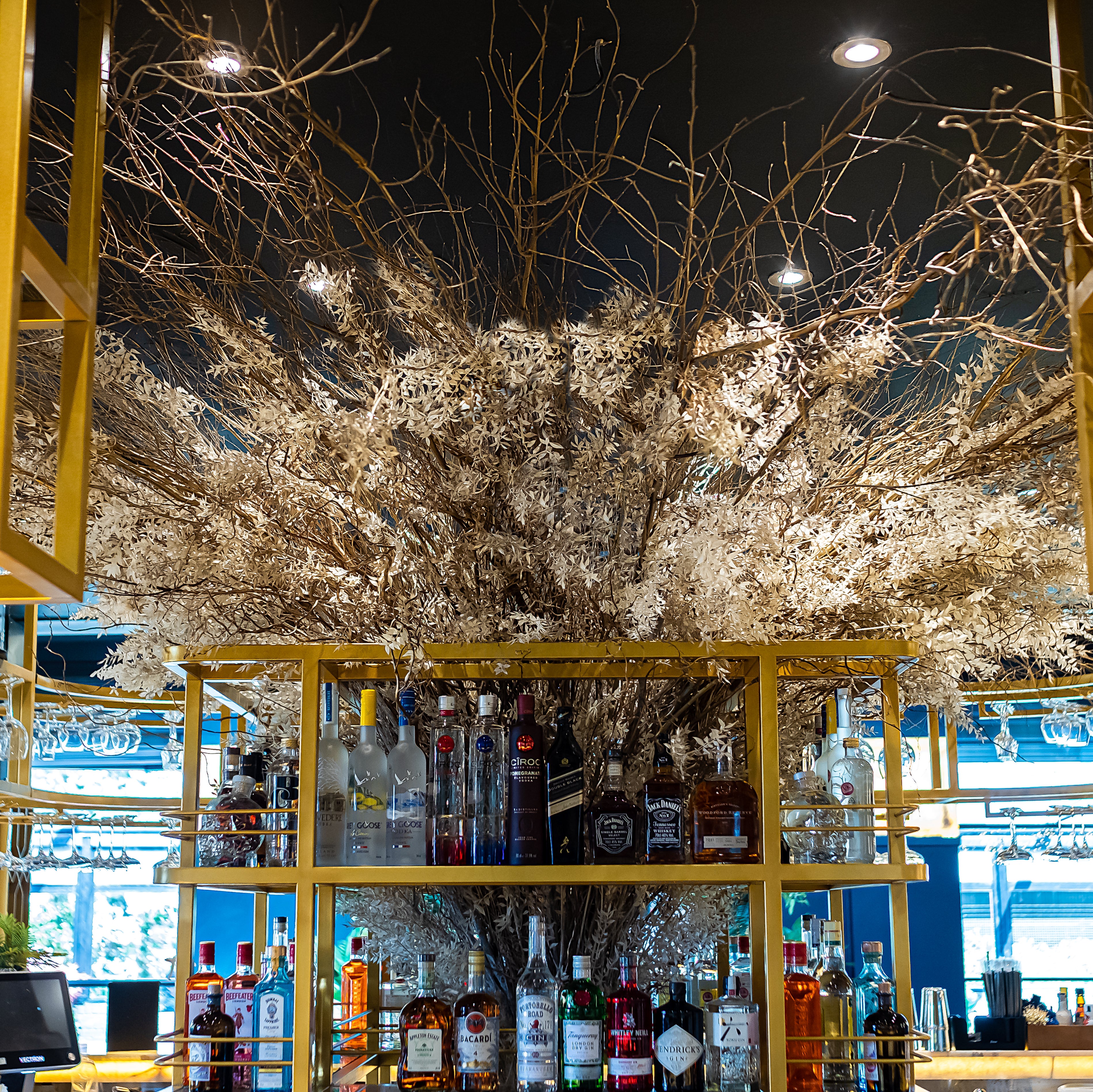 An intricate and expansive preserved floral tree installation by Amaranté London, featuring delicate branches and pale foliage, creates a dramatic centrepiece above the bar area of Nova London, a fashion wholesaler. The natural textures of the tree contrast with the neatly arranged bottles of spirits on the modern, yellow metal shelves below.