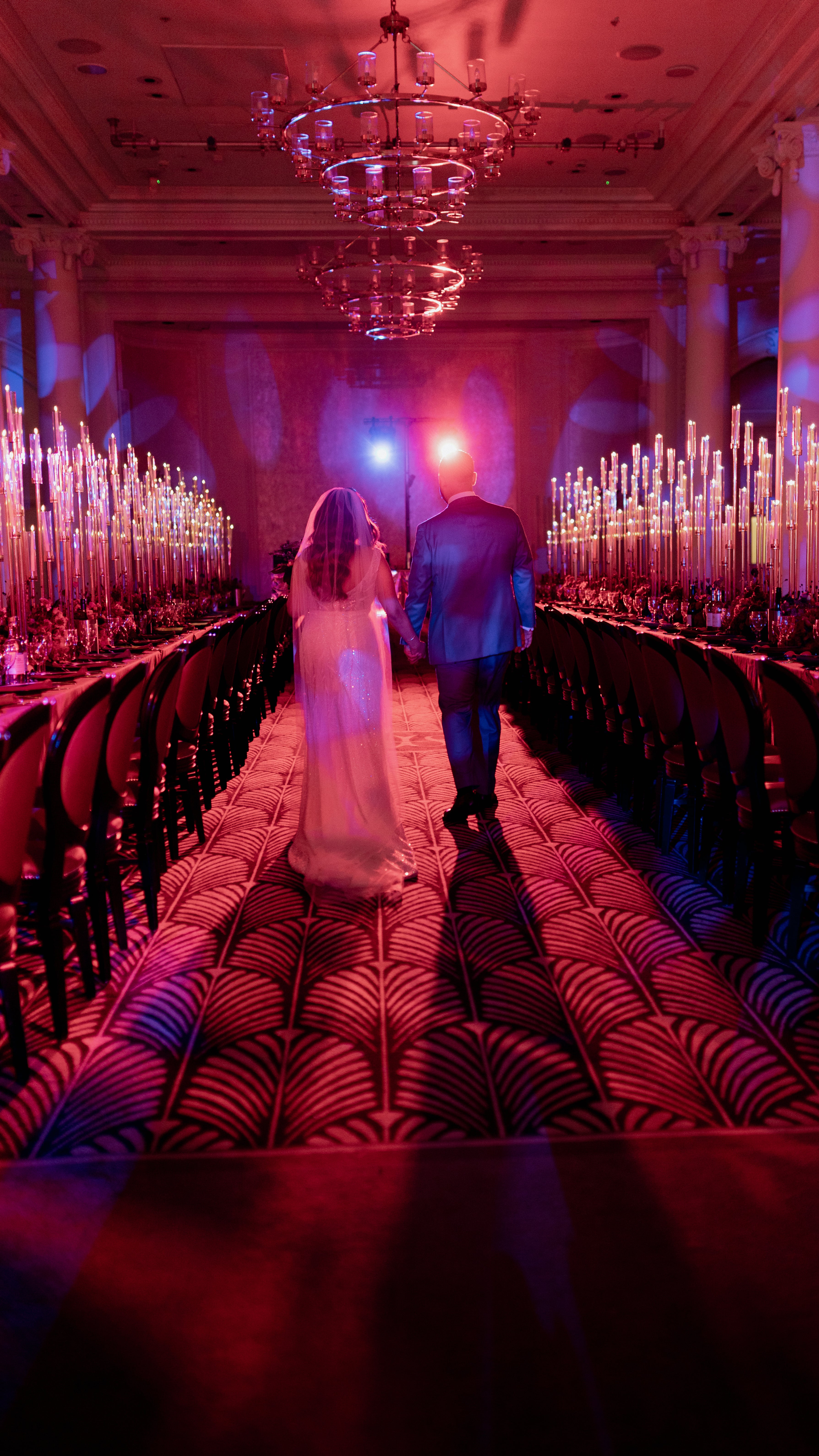 Bride and groom walking down a candlelit aisle at the Waldorf Hilton with hundreds of tall taper candles, low red floral arrangements, and pink atmospheric lighting - Amaranté London.