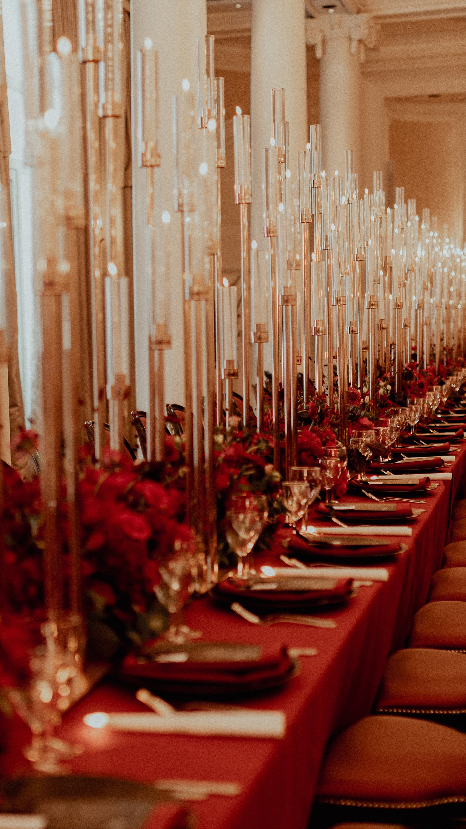 Striking forest of tall taper candles in gold holders with low red floral arrangements creating dramatic candlelit tablescape at Waldorf Hilton banquet reception - Amaranté London.