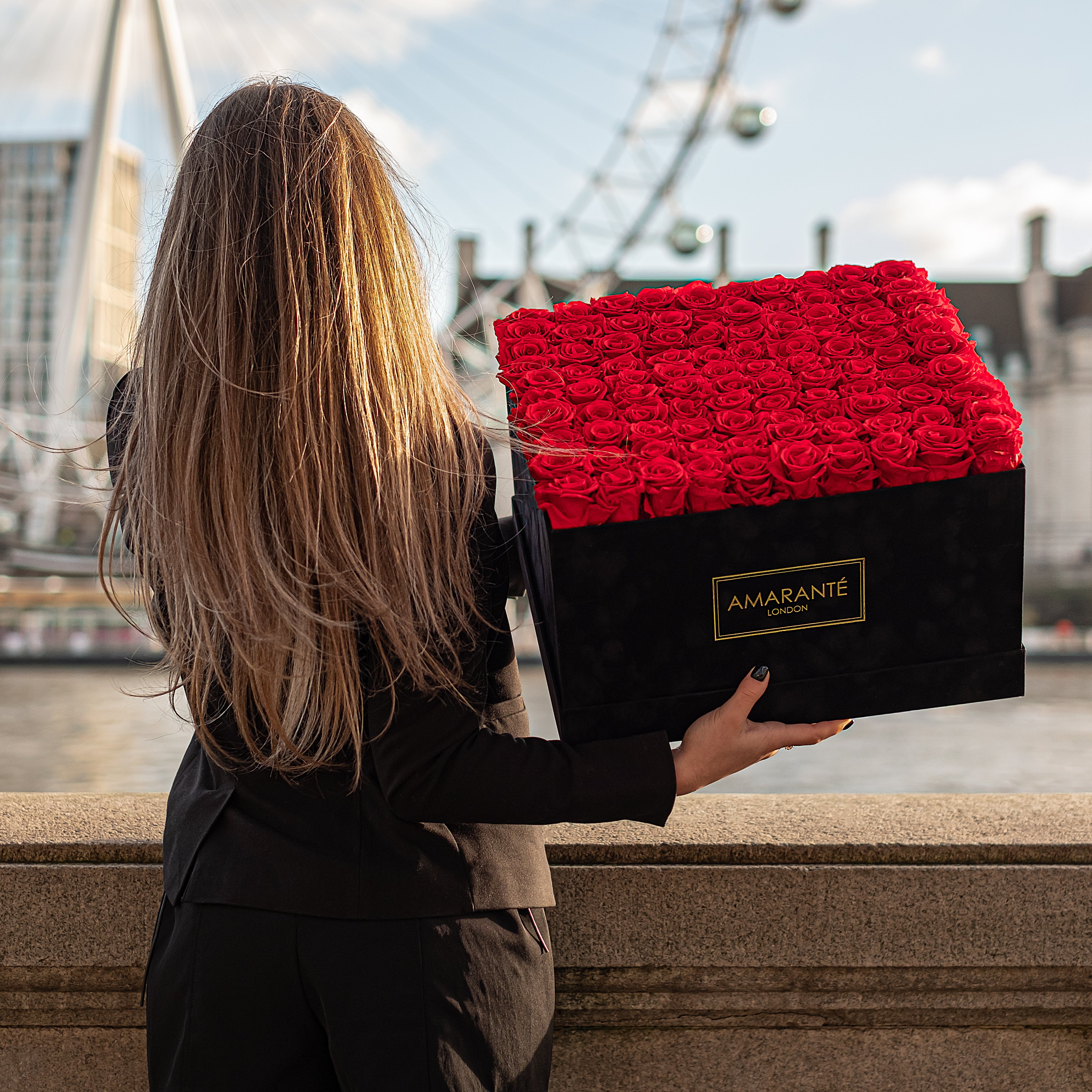 A woman elegantly dressed with long blond hair, seen from behind, holding a large square black suede box filled with lush red infinity roses, with the London Eye in the background - Amaranté London.