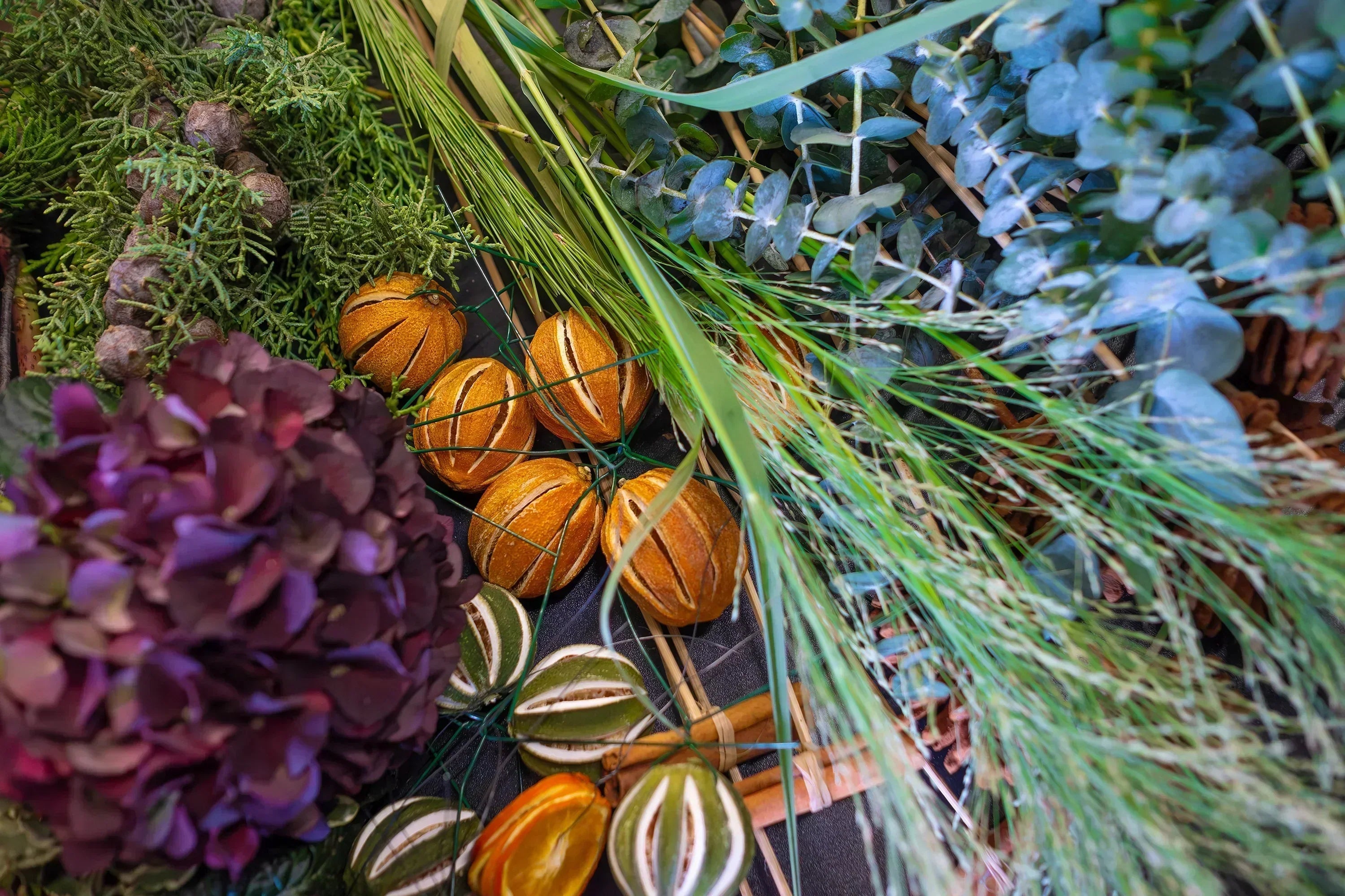 Close-up of luxury Christmas wreath materials including dried orange slices, eucalyptus, burgundy hydrangeas, fresh pine, and cinnamon bundles arranged on dark surface - Amarante London