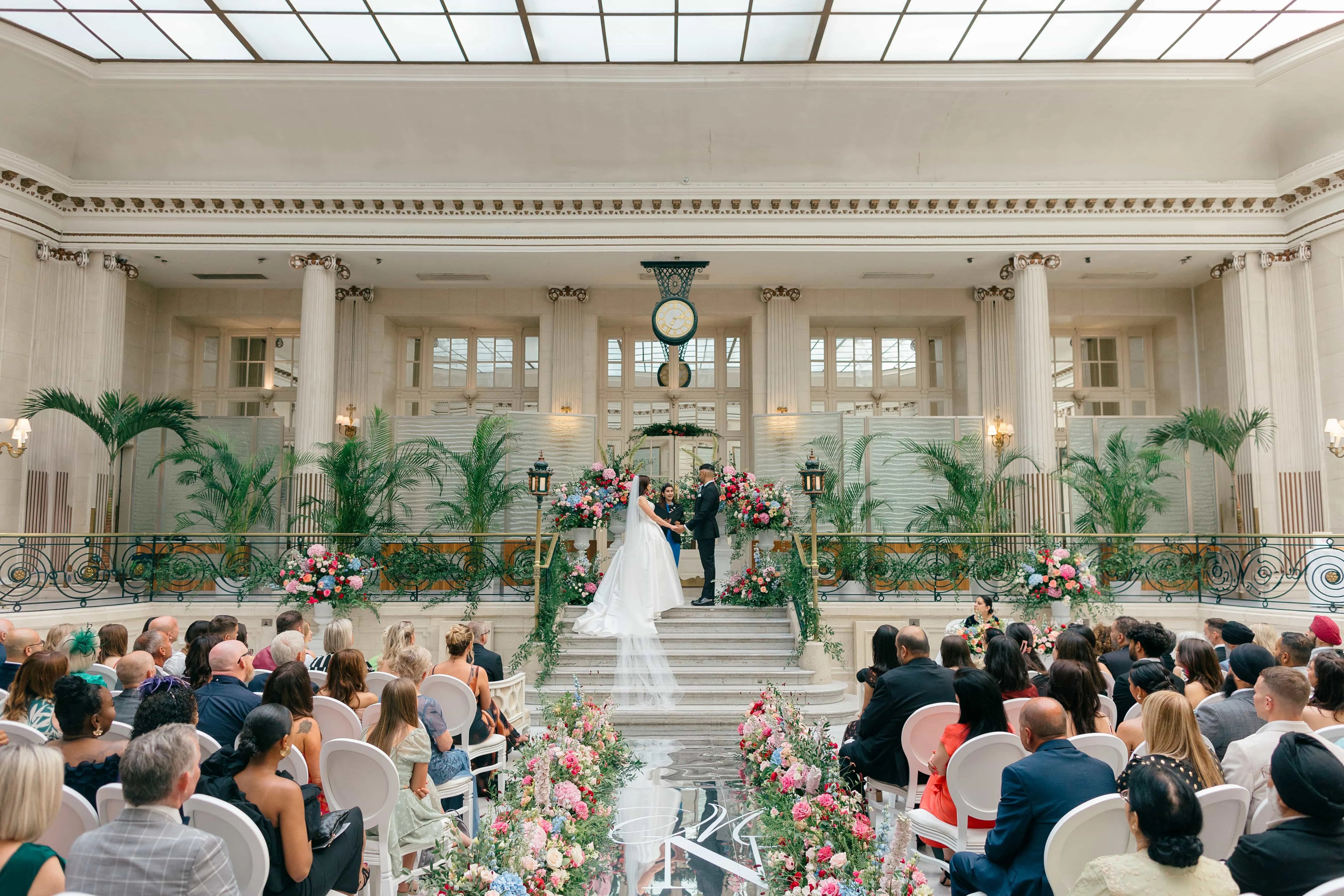 Wedding ceremony at Waldorf Hilton with bride and groom on grand staircase, floral arrangements, palm trees, and guests in elegant ballroom with classical columns - floral design by Amaranté London