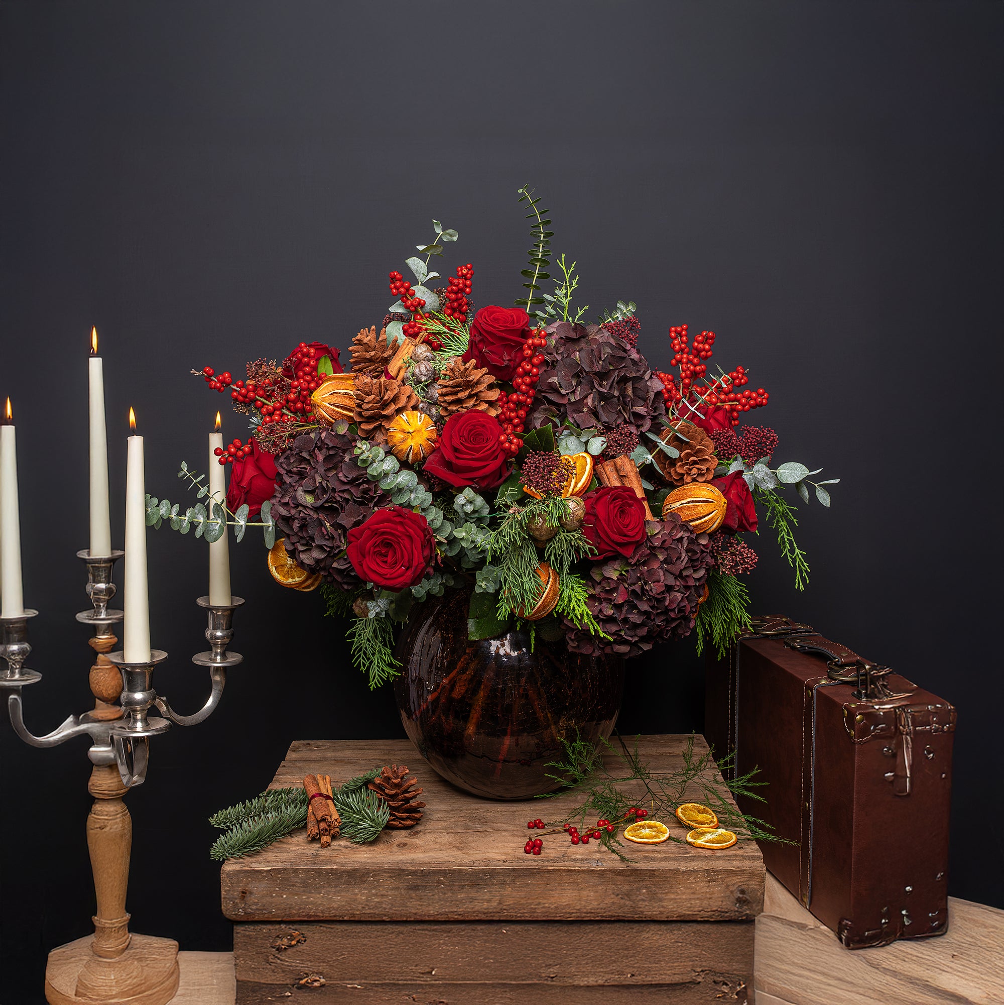 Luxury Christmas flower arrangement featuring deep red roses, dark burgundy hydrangeas, and bright red berry stems with eucalyptus foliage and fresh evergreen sprigs, accented with cinnamon sticks tied in twine and dried orange slices, arranged in a dark glass vase on a wooden surface with silver candlesticks and vintage styling - Amaranté London