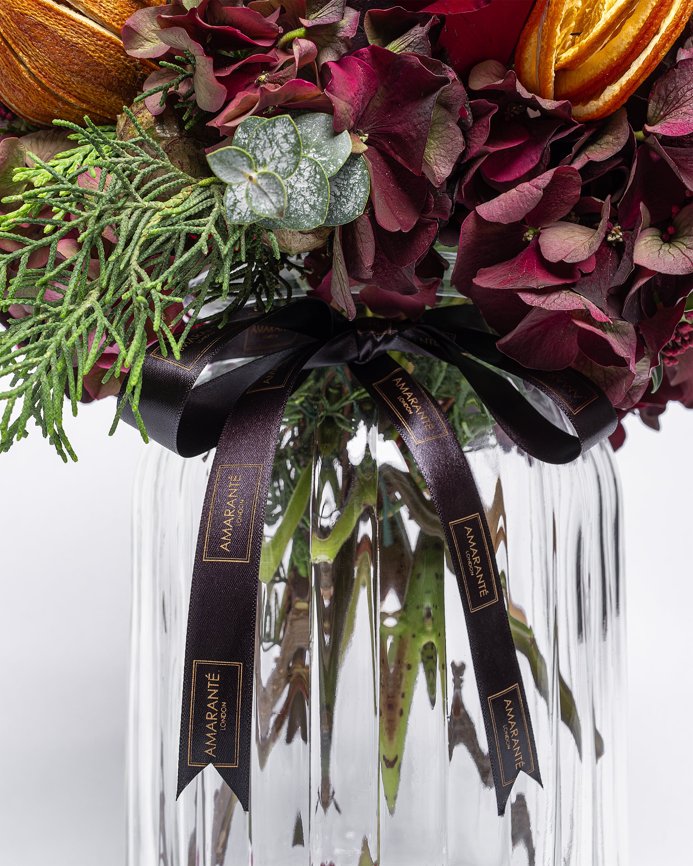 Bouquet of flowers with greenery and oranges in a clear vase, featuring a black ribbon with 'Mamie' branding.