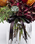 Bouquet of flowers with greenery and oranges in a clear vase, featuring a black ribbon with 'Mamie' branding.