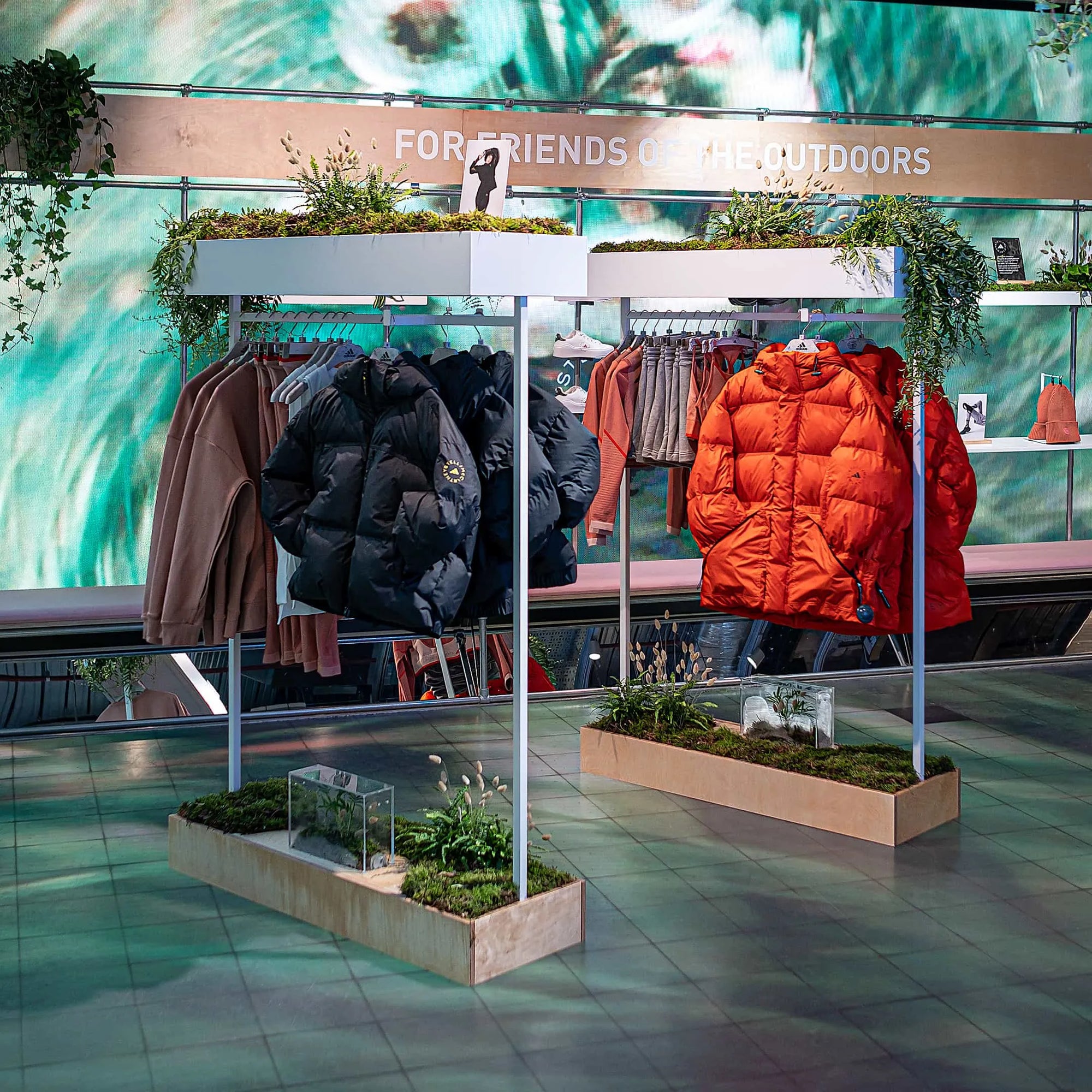 Amaranté London's exquisite event floristry frames an Adidas x Stella McCartney aSMC collection display, showcasing winter jackets above terrarium-like planters. The greenery imbues the space with vitality, highlighting the collection's outdoor-friendly ethos.