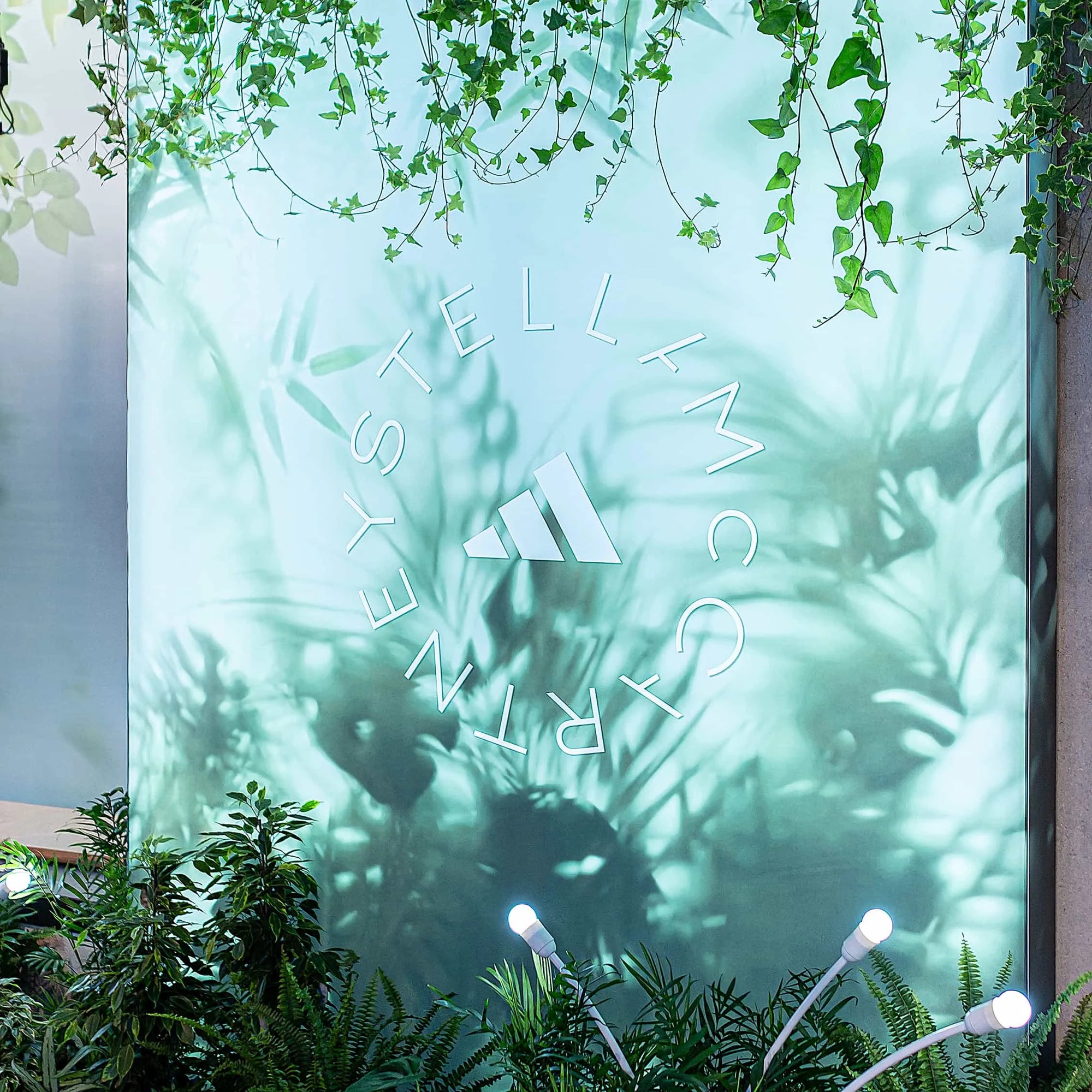 In this image, the signature Adidas x Stella McCartney aSMC logo is accentuated by a canopy of delicate ivy and verdant plants, artfully arranged by Event Florist Amaranté London. A botanical backdrop was implemented for this strategic retail space, setting a natural ambience to the high-end fashion setting.