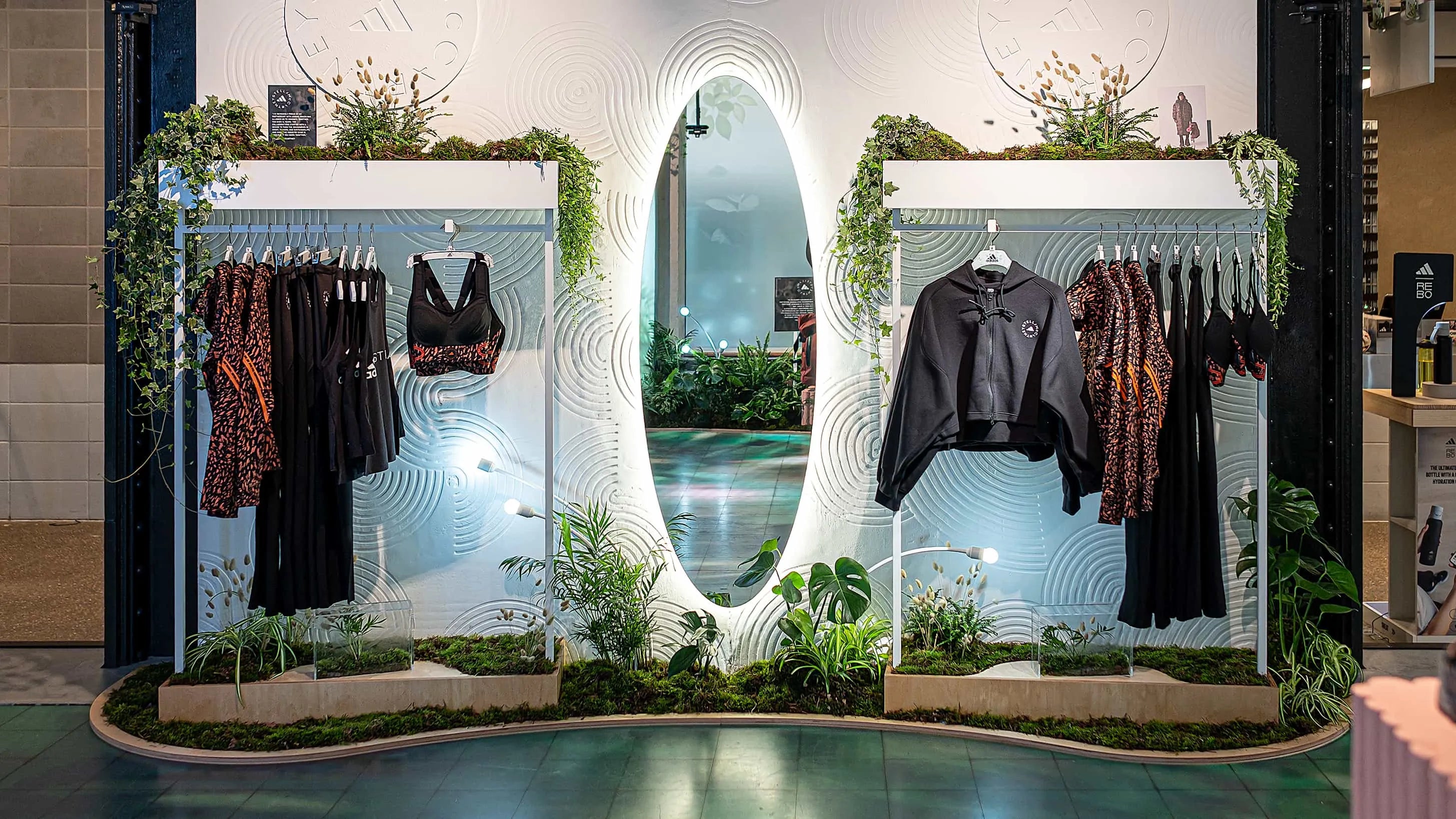 This Amaranté London-designed setup for the Adidas x Stella McCartney aSMC collection launch pairs athletic wear with carefully chosen plants and greenery. The archway, bordered by beds of lush foliage, frames the stylish apparel, creating a sophisticated, relaxing and sustainable retail space at the Adidas x Stella McCartney aSMC collection launch.