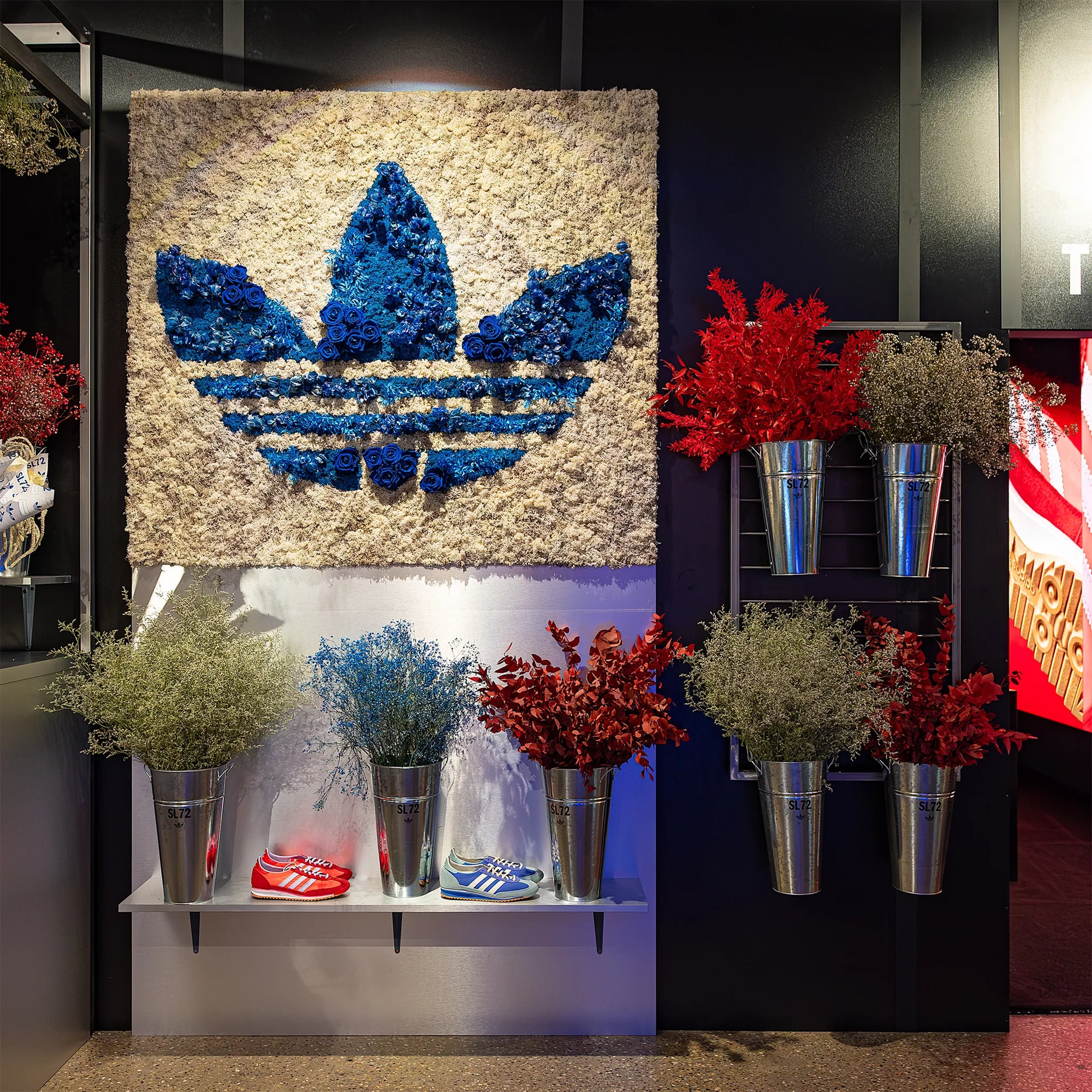 A floral arrangement designed by Amaranté London on display at the Adidas Oxford Street Store in London for the SL72 collection launch. The image shows a blue Adidas logo made from preserved flowers on a textured background with plants in metal planters.