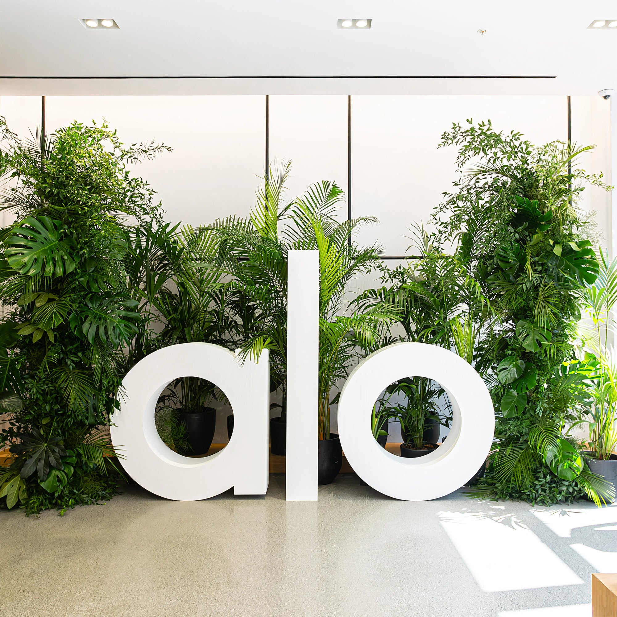In this image, large custom-designed white 'Alo' letters are surrounded by lush tropical plants, including Monstera and Kentia palms, at the Alo Regent Street Store Grand Opening, created by Amaranté London. The greenery emphasises the store's luxurious, wellness-focused theme. Amaranté London, Event Florist