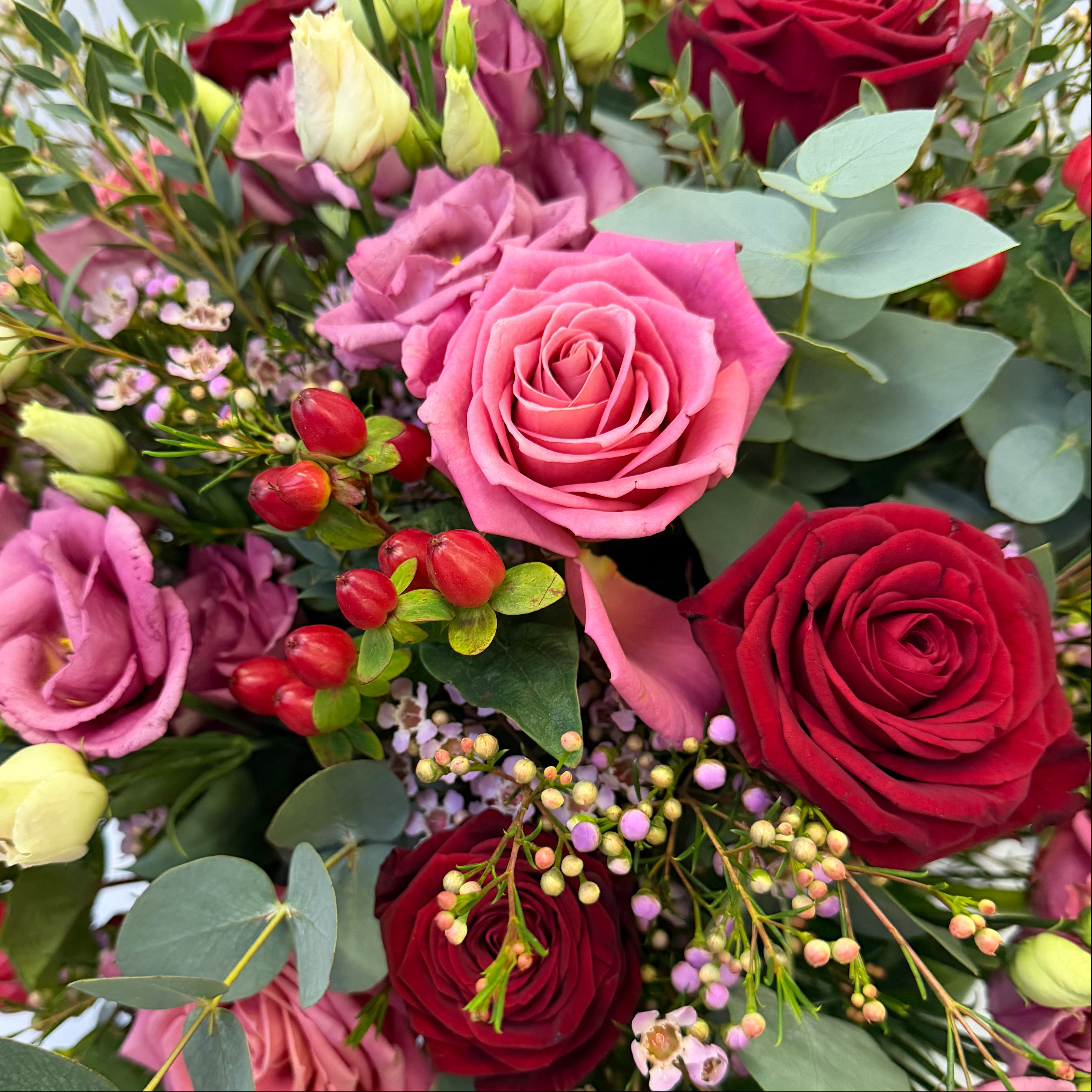 Close-up detail of Aurora bouquet showing velvety red roses, vibrant pink ranunculus, pale pink lisianthus blooms, waxflower clusters, orange hypericum berries, and silver eucalyptus leaves - Amaranté London
