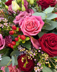 Close-up detail of Aurora bouquet showing velvety red roses, vibrant pink ranunculus, pale pink lisianthus blooms, waxflower clusters, orange hypericum berries, and silver eucalyptus leaves - Amaranté London