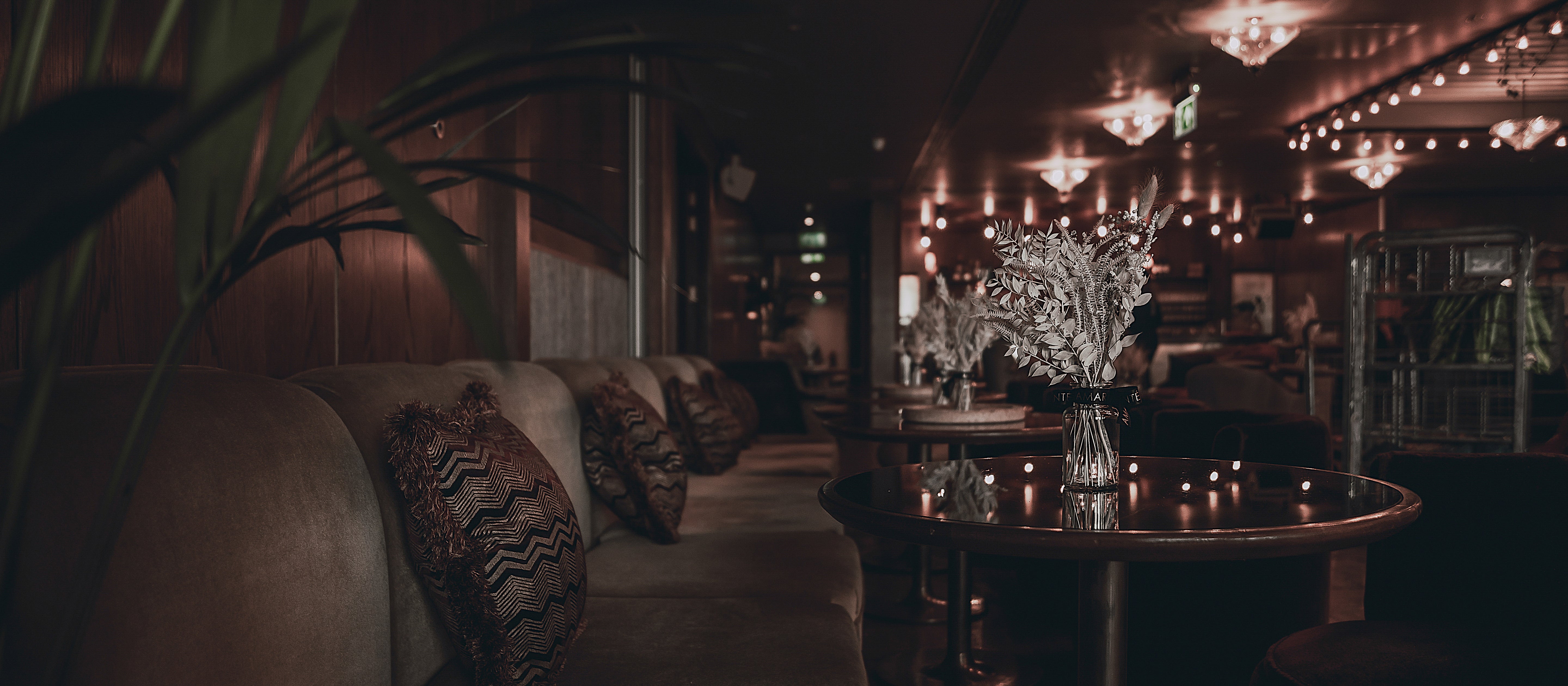 A panoramic view of White City House's sophisticated lounge area, adorned with Amaranté's bespoke floral arrangements for events, featuring a series of elegant vases filled with silvery botanicals on round tables, enhancing the venue's warm, inviting ambience with a touch of botanical chic.