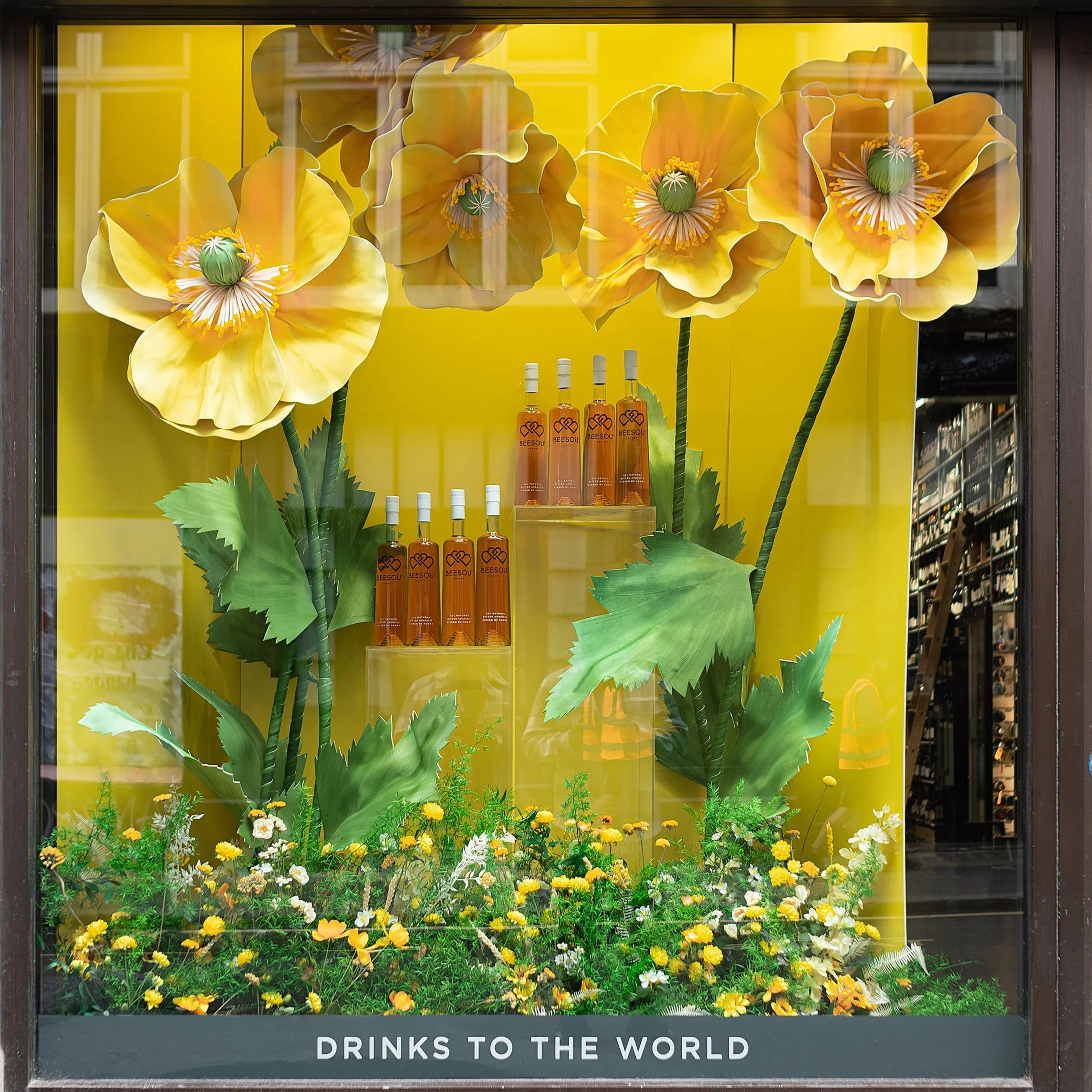 Close-up of Beesou bottles displayed within Amaranté London's bespoke floral window installation at Amathus, Wardour Street, London. The display's yellow poppies and lush greenery highlight the aperitif brand and enhance the visual appeal of the storefront.