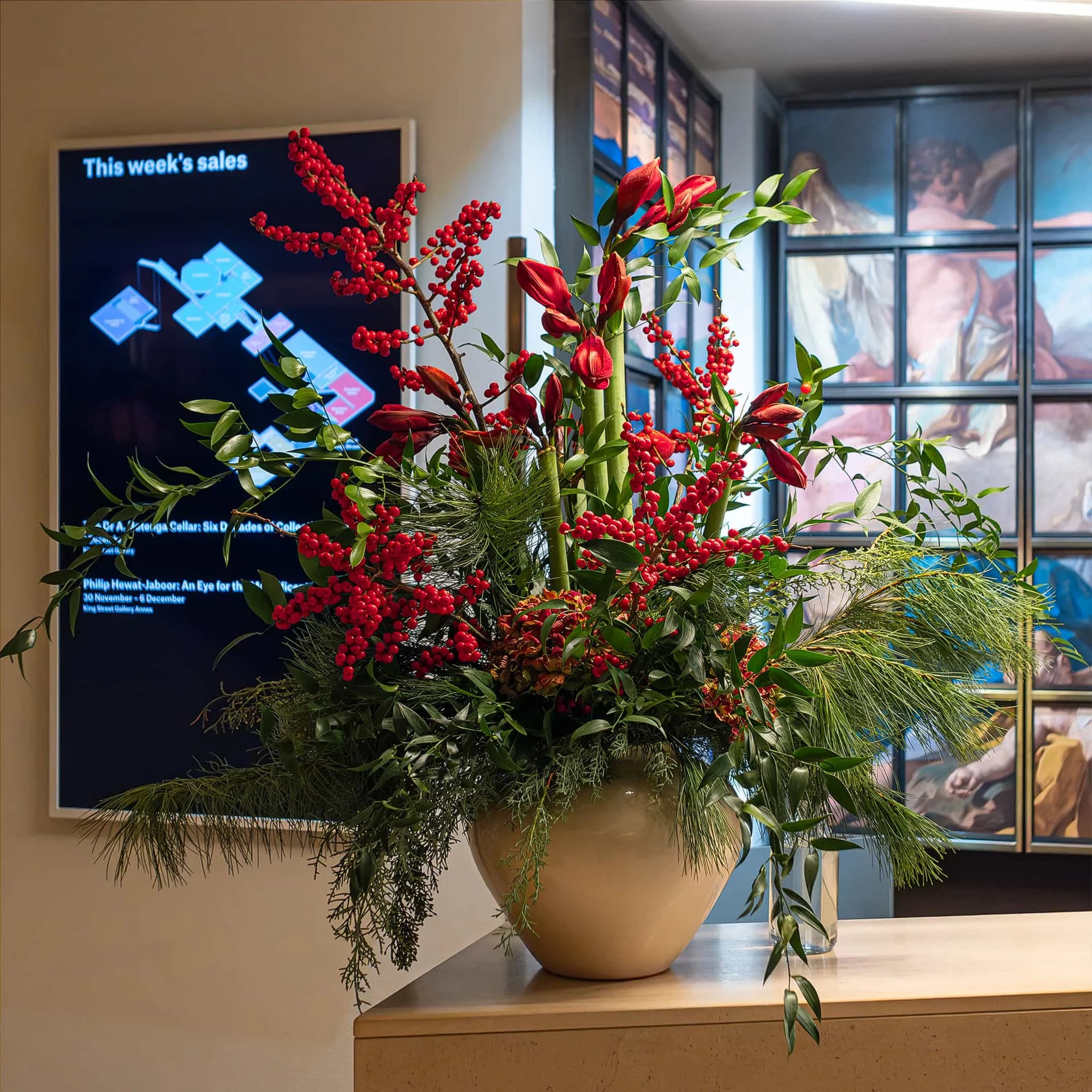 A Bespoke Christmas Bouquet at Christie's reception desk features rich red Alstroemeria flowers, vivid Ilex berries, an assortment of lush greenery, and pine needles, beautifully displayed in a modern beige vase.