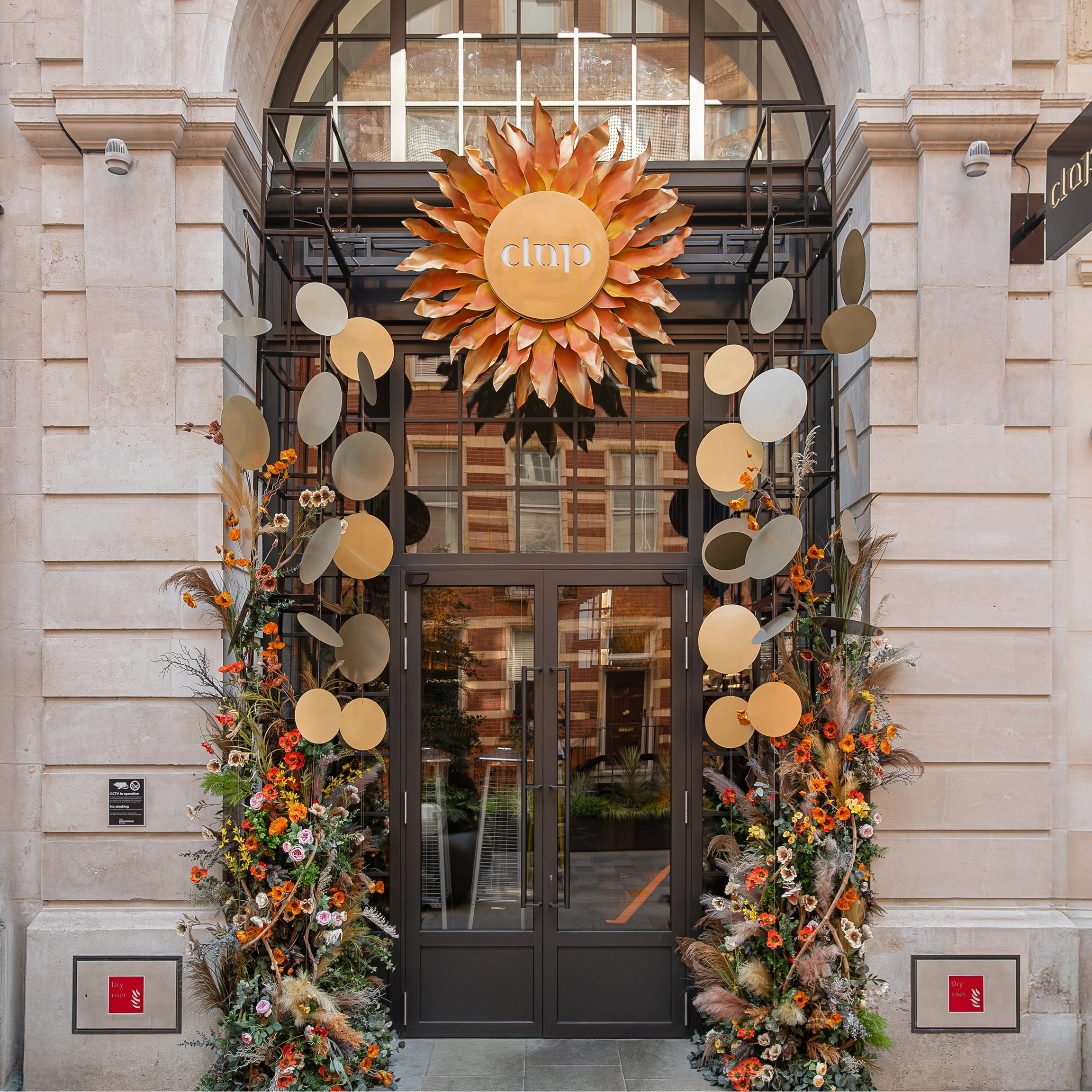Summer solstice floral entrance installation with sunburst centrepiece, dried botanicals and seasonal flowers, created for Clap Restaurant Knightsbridge by Amarante London