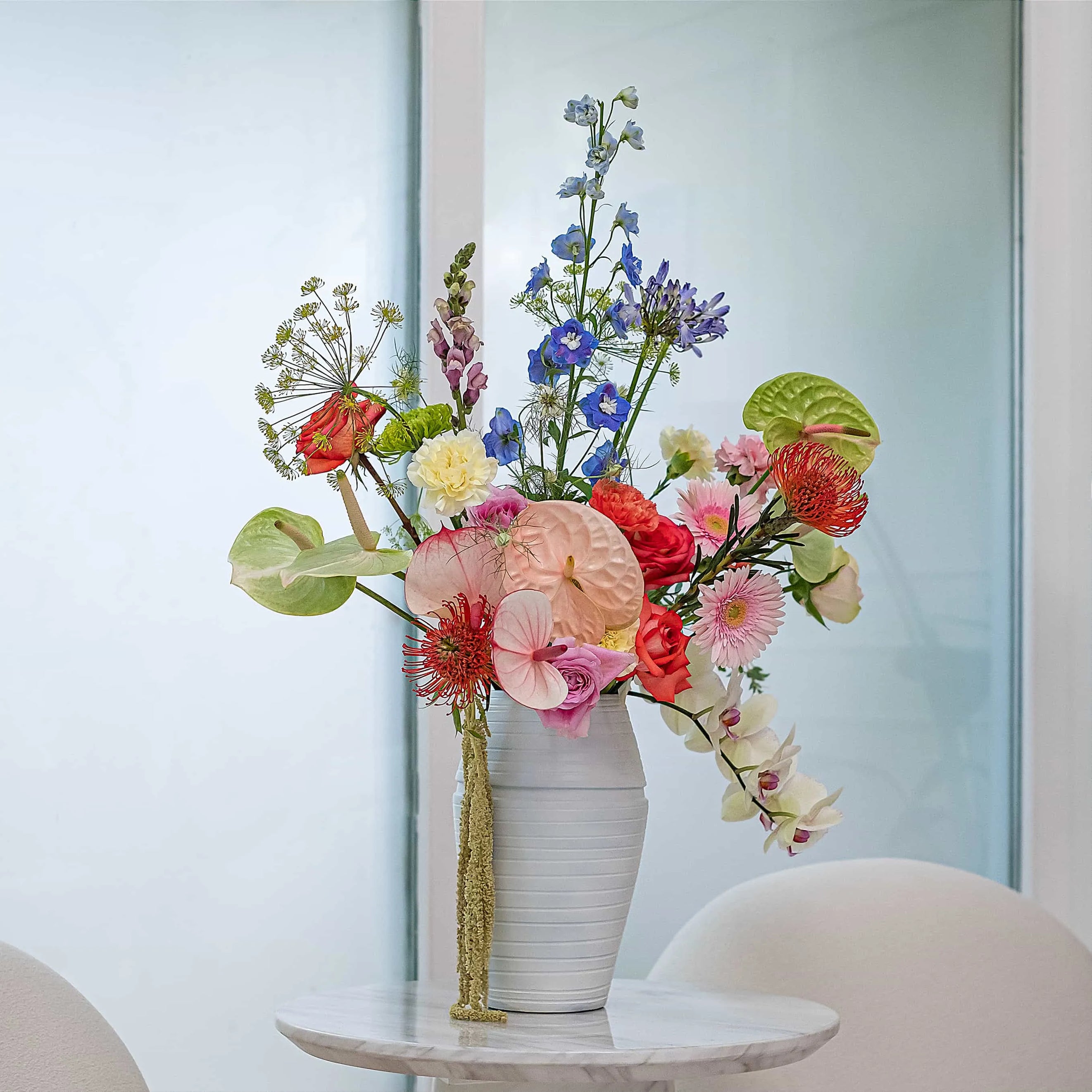 Weekly office flower arrangement featuring anthurium, blue delphiniums, orchids and mixed seasonal blooms in a white ceramic vase, London corporate flower contract by Amarante London