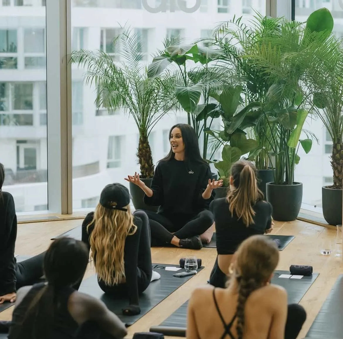 A Yoga class in a well-lit, spacious room with floor-to-ceiling windows. A selection of large, potted plants in the background fills the space with a sense of calm and a connection to nature. Plant rental services by Amaranté London are designed to enhance the wellness experience for corporate or business clients.