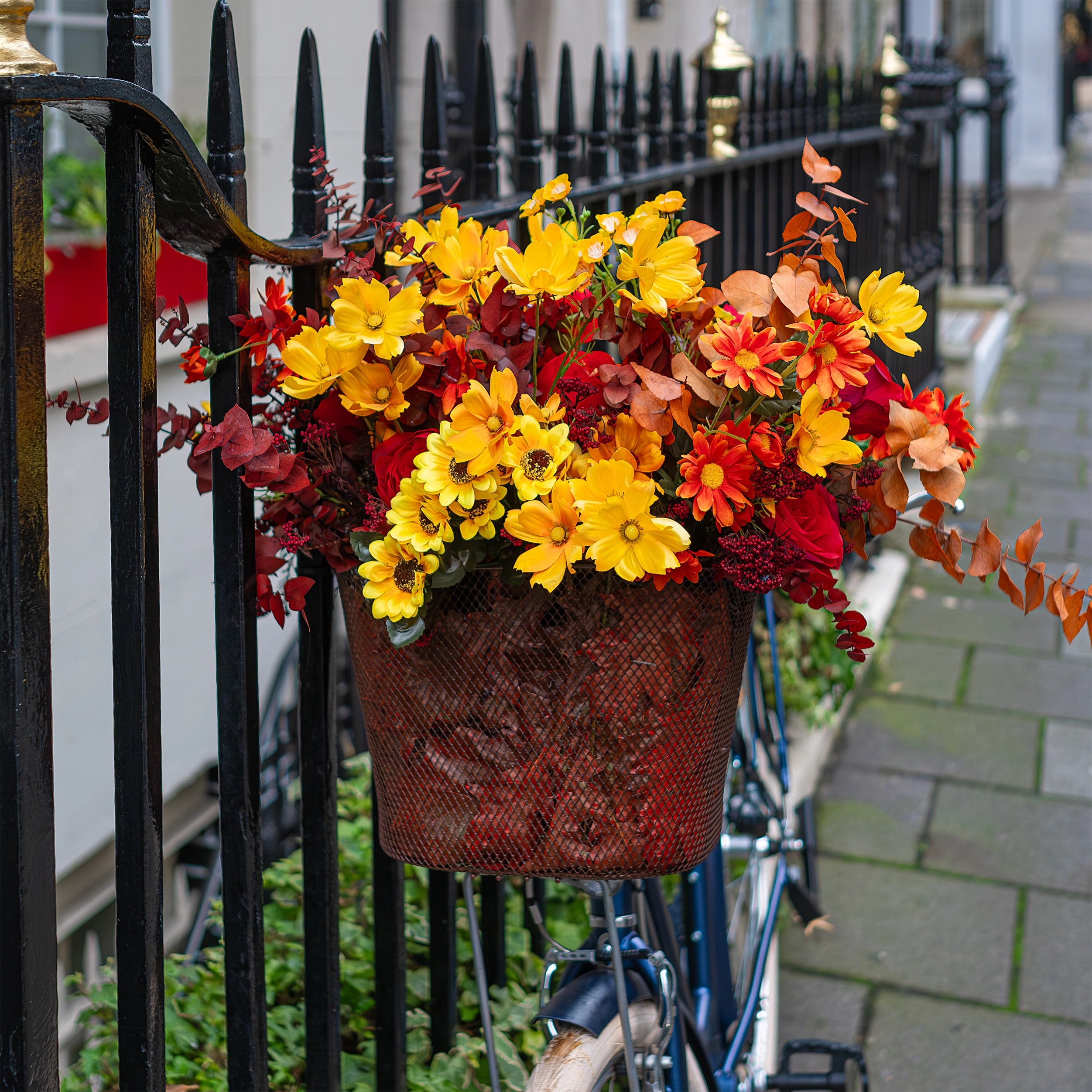 A whimsical floral basket on a vintage blue bicycle for Crown London’s autumn display. The basket features a vibrant mix of yellow cosmos, red roses, and orange marigolds, creating a lively contrast that complements the venue’s seasonal decor with a playful and elegant touch - Floral design by Amaranté London Event Florist  