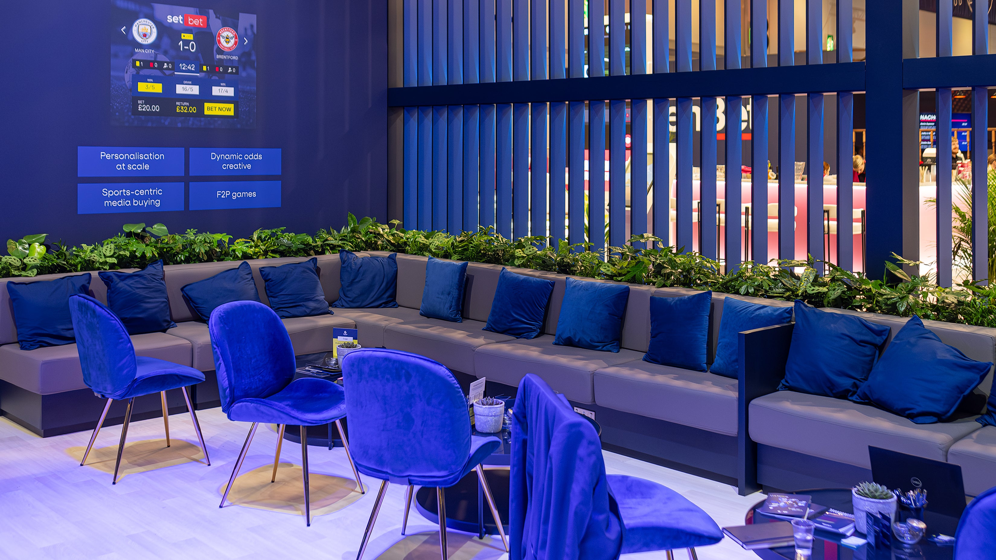 At ICE London Exhibition, the Genius Sports lounge area is elegantly decorated with plush blue cushions and a greenery wall designed and produced by Amaranté London Event Florist. The intense green plants were specifically chosen to complement the digital displays showcasing dynamic sports betting odds.