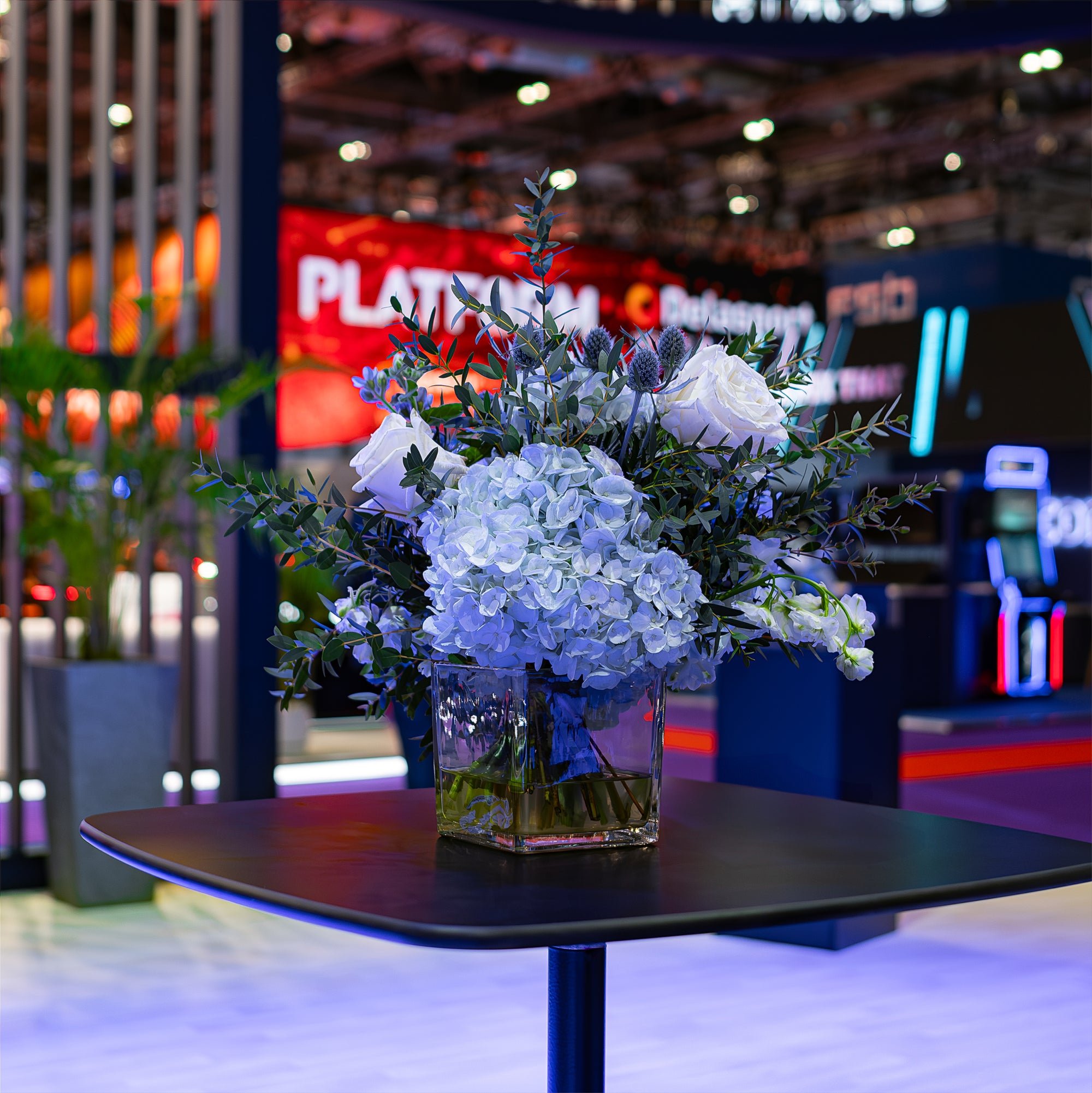 An exquisite arrangement of pale blue hydrangeas and pristine white roses, displayed at ICE London Exhibition, accents a corporate networking area, infusing elegance into the vibrant atmosphere of this corporate event.