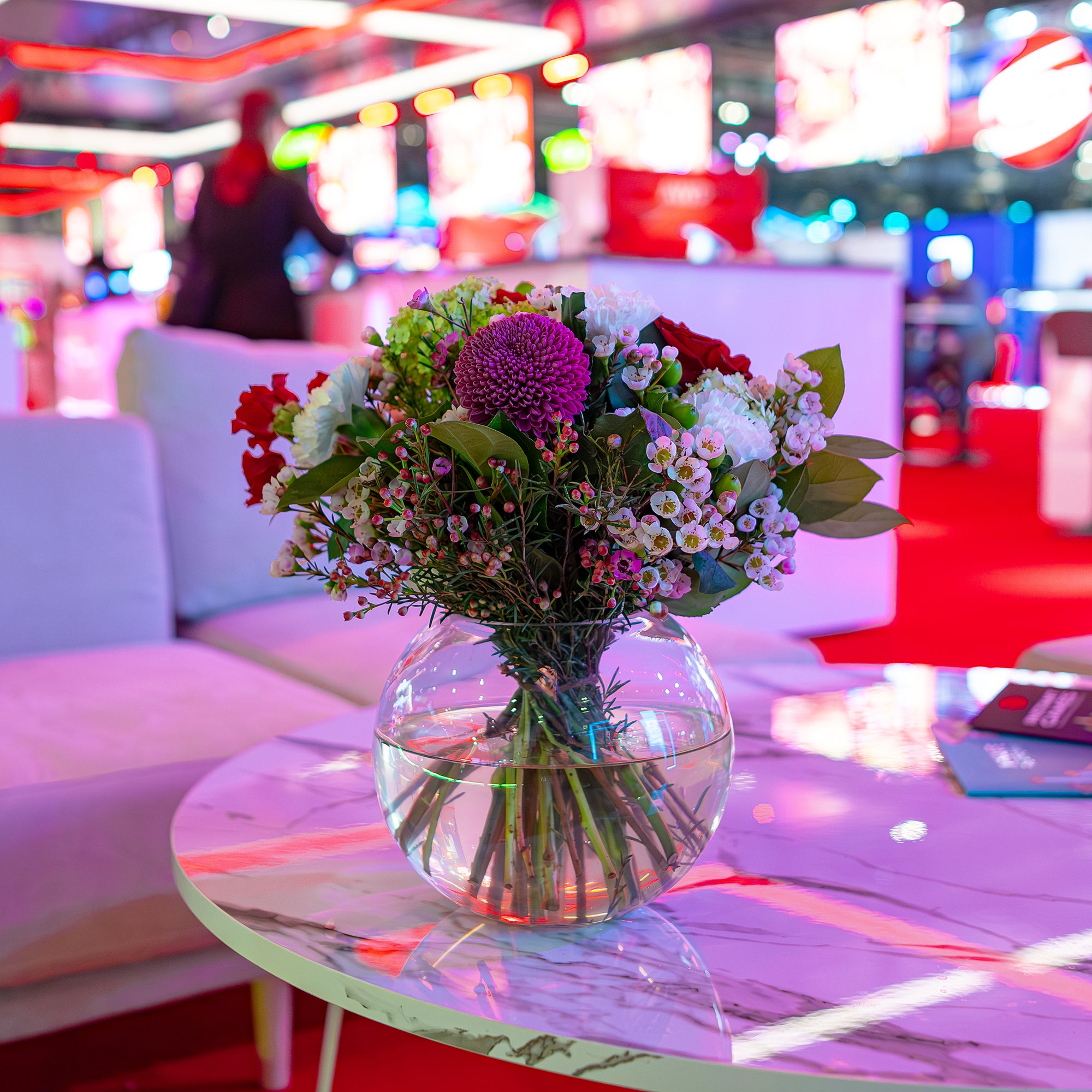 A vibrant floral bespoke centrepiece designed and created by Amaranté London features rich purples and reds. The bouquet sits elegantly on a table at ICE London Exhibition, creating an inviting atmosphere amidst the dynamic energy of this dynamic Corporate Event.