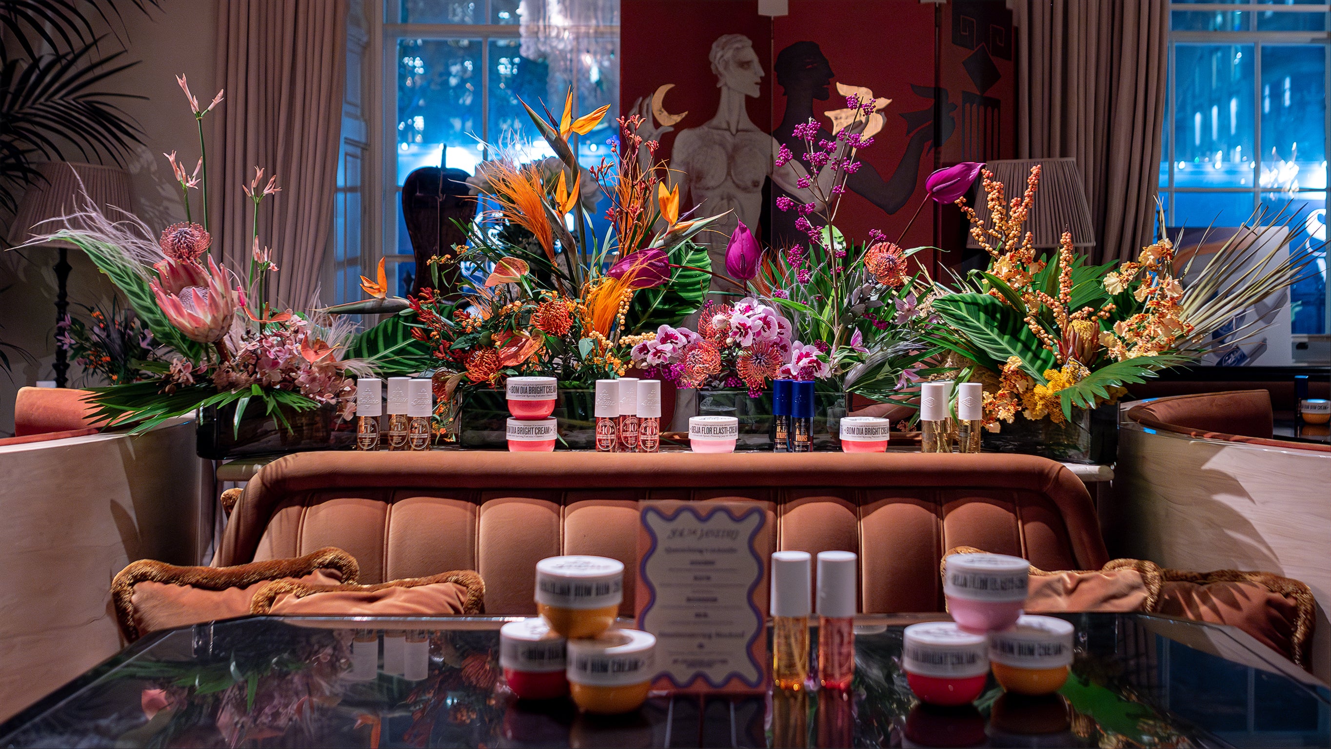 A lavish array of Sol de Janeiro's beauty products elegantly presented on a grand table amid Amaranté London's striking floral arrangements, with a backdrop of chic interior decor at Maine Mayfair restaurant.
