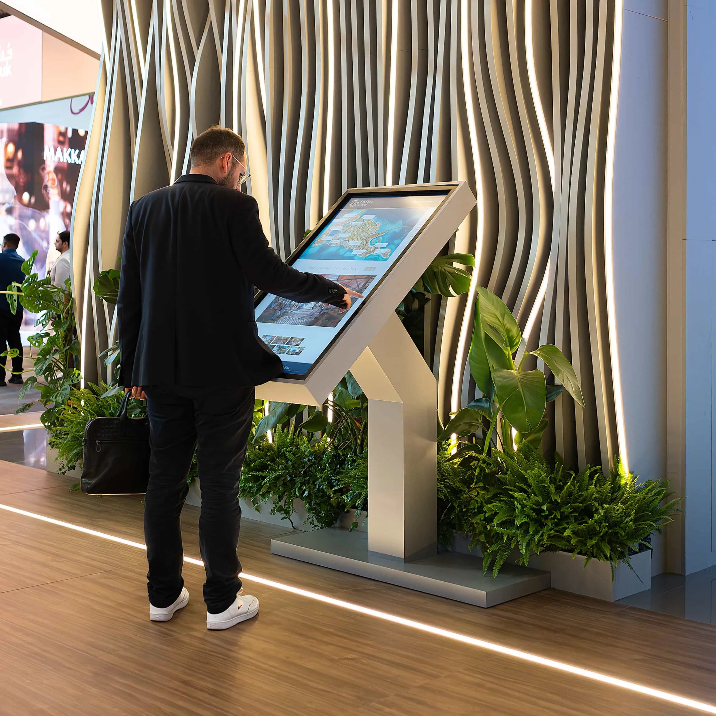A modern interactive kiosk with a man in a black jacket and jeans browsing through a digital display stands prominently in a corporate event. Surrounding the kiosk are lush green ferns. Intersperse between the sleek, vertical, wavy lines of the sculptural backdrop illuminated by warm, ambient lighting stand large, vibrant green plants hired for the exhibition, curated by Amaranté London event florist. These plants for the exhibition create a refreshing and engaging atmosphere within the business setting.