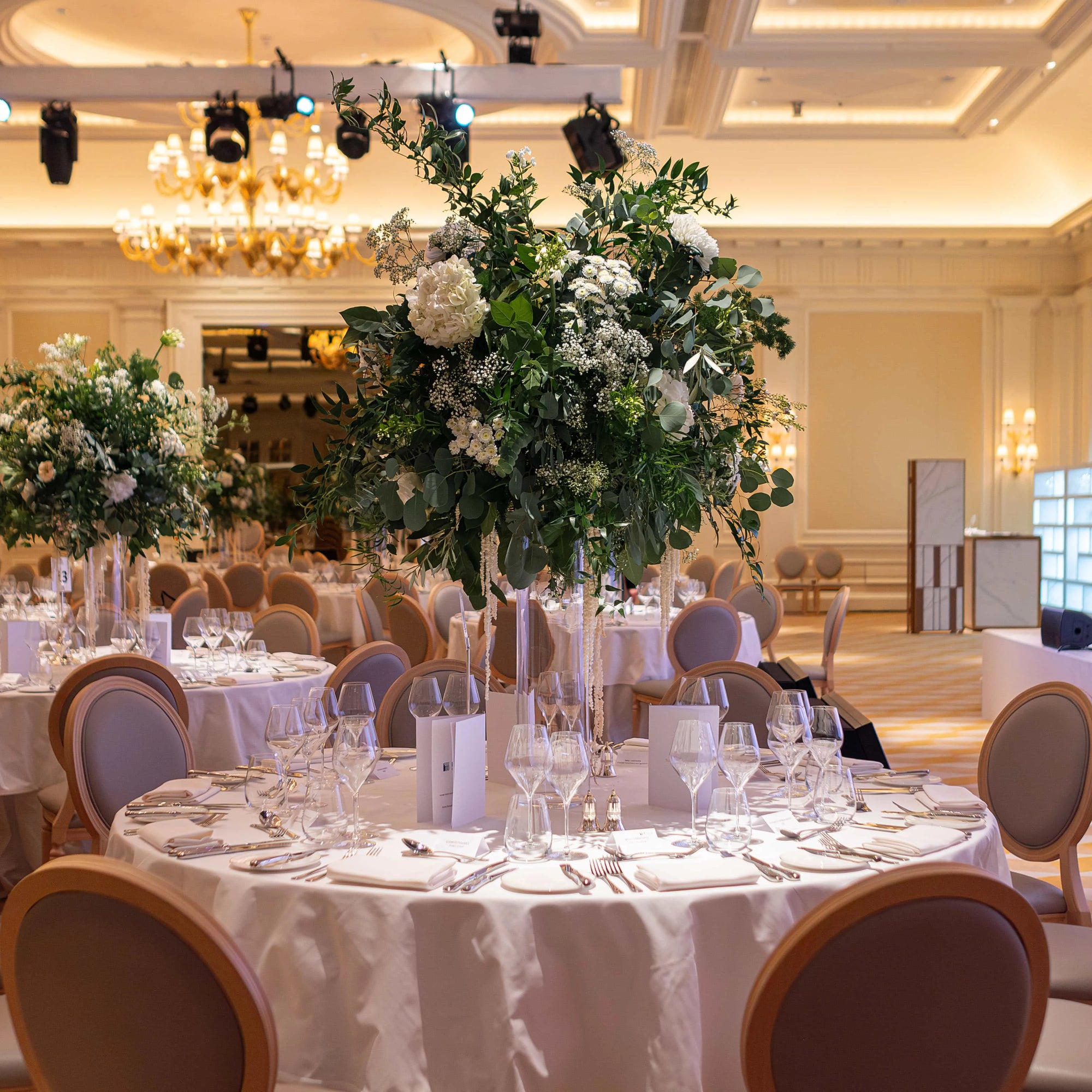 This is a detailed view of a round table at the FTI Consulting corporate event adorned with a stunning bespoke floral centrepiece by Amaranté London. The centrepiece features lush greenery and pristine white flowers.