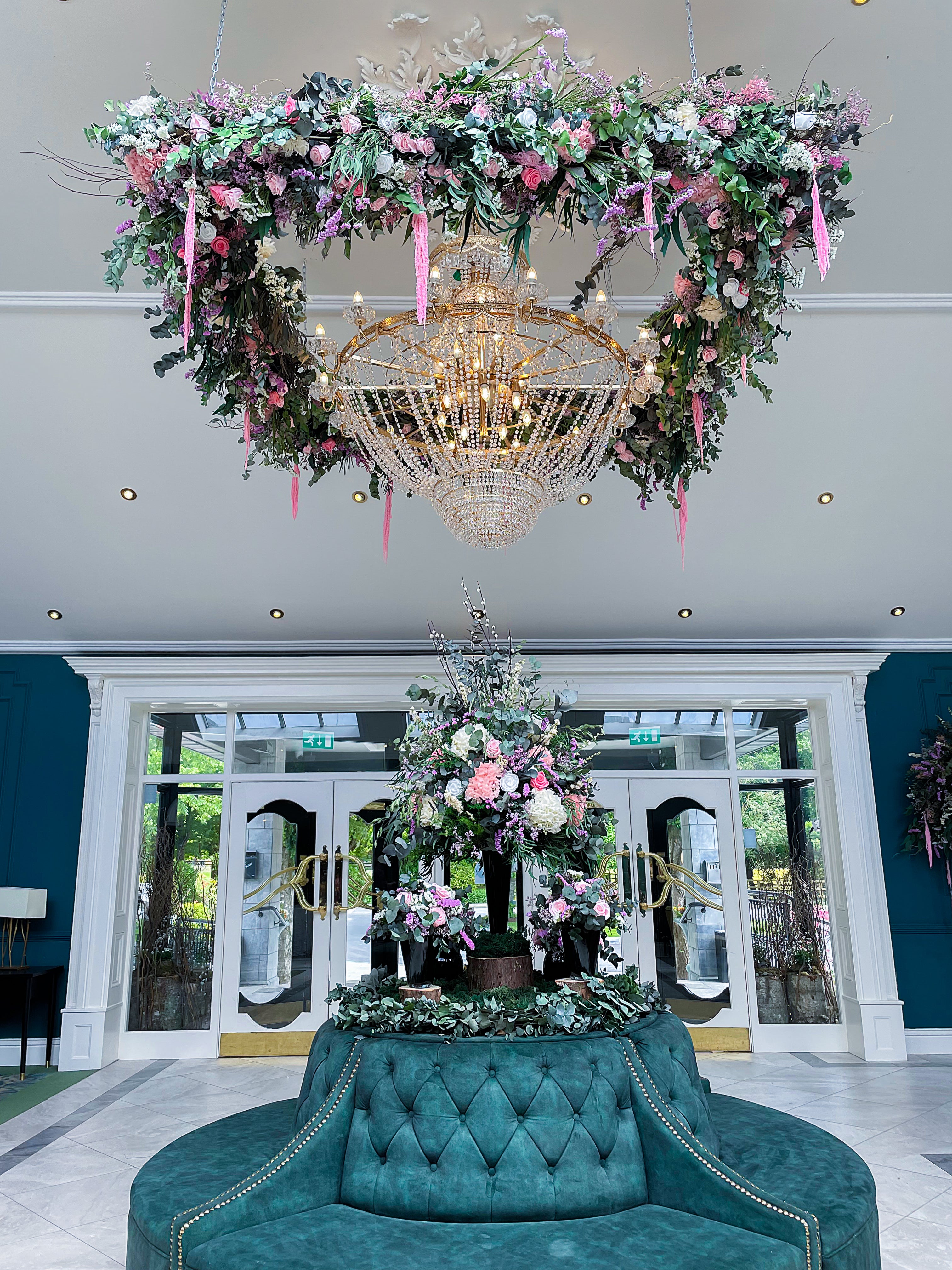 A luxurious floral installation cascades from an ornate chandelier in the lobby of Fitzgerald Woodlands House Hotel & Spa in Ireland, featuring a bespoke floral installation with various flowers in shades of pink, lavender, and cream amid lush greenery. This elaborate display hangs above a rich teal, tufted circular couch, contributing to the warm and welcoming atmosphere of the hotel.