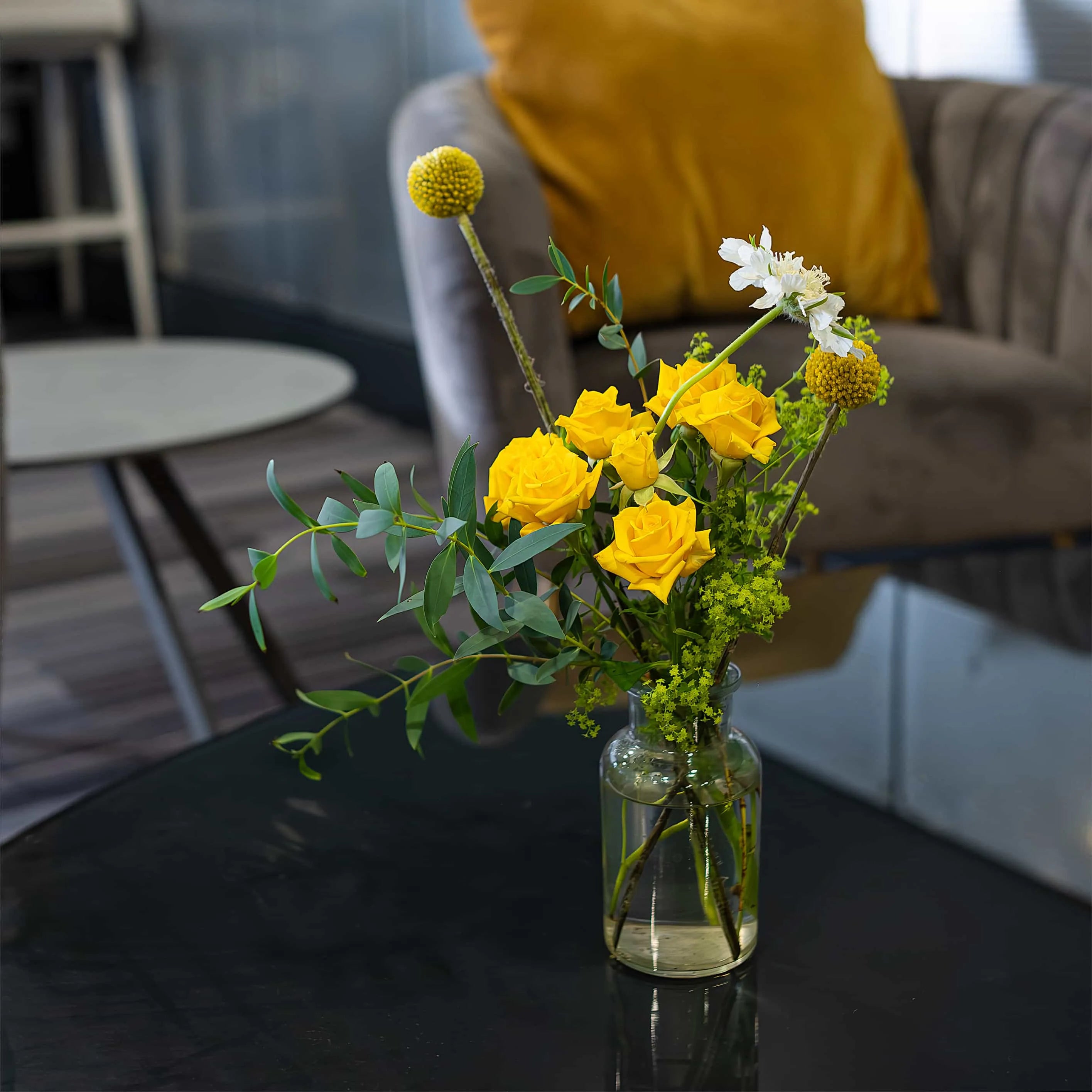 A bright and elegant floral display featuring vivid yellow roses and spherical green blooms arranged in a clear glass vase on a dark table adds colour and freshness to a lounge area with a modern look.