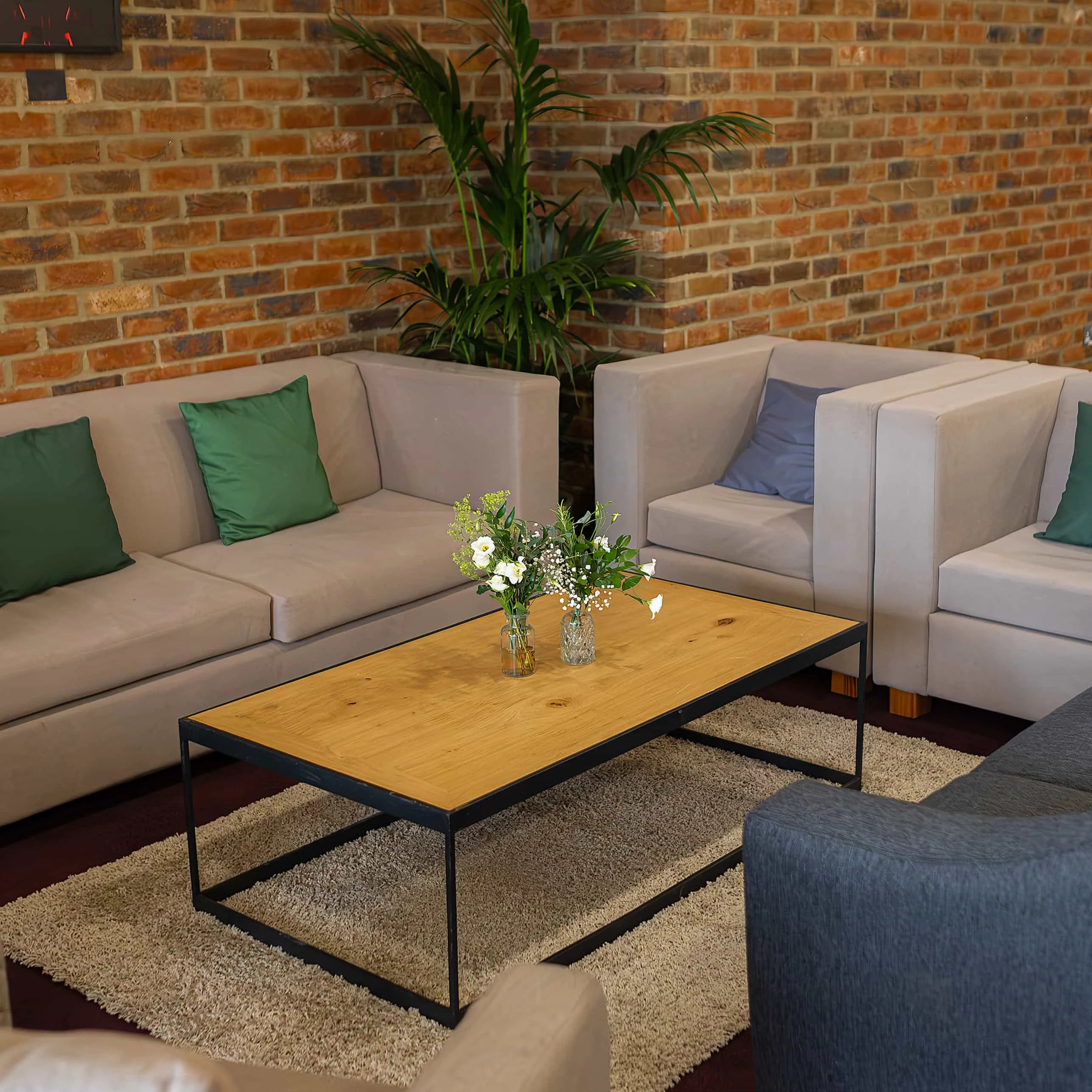 An elegant floral arrangement on a lounge coffee table with green and white flowers in clear vases enhances a relaxed meeting environment with a brick wall backdrop and plush seating - Amaranté London 