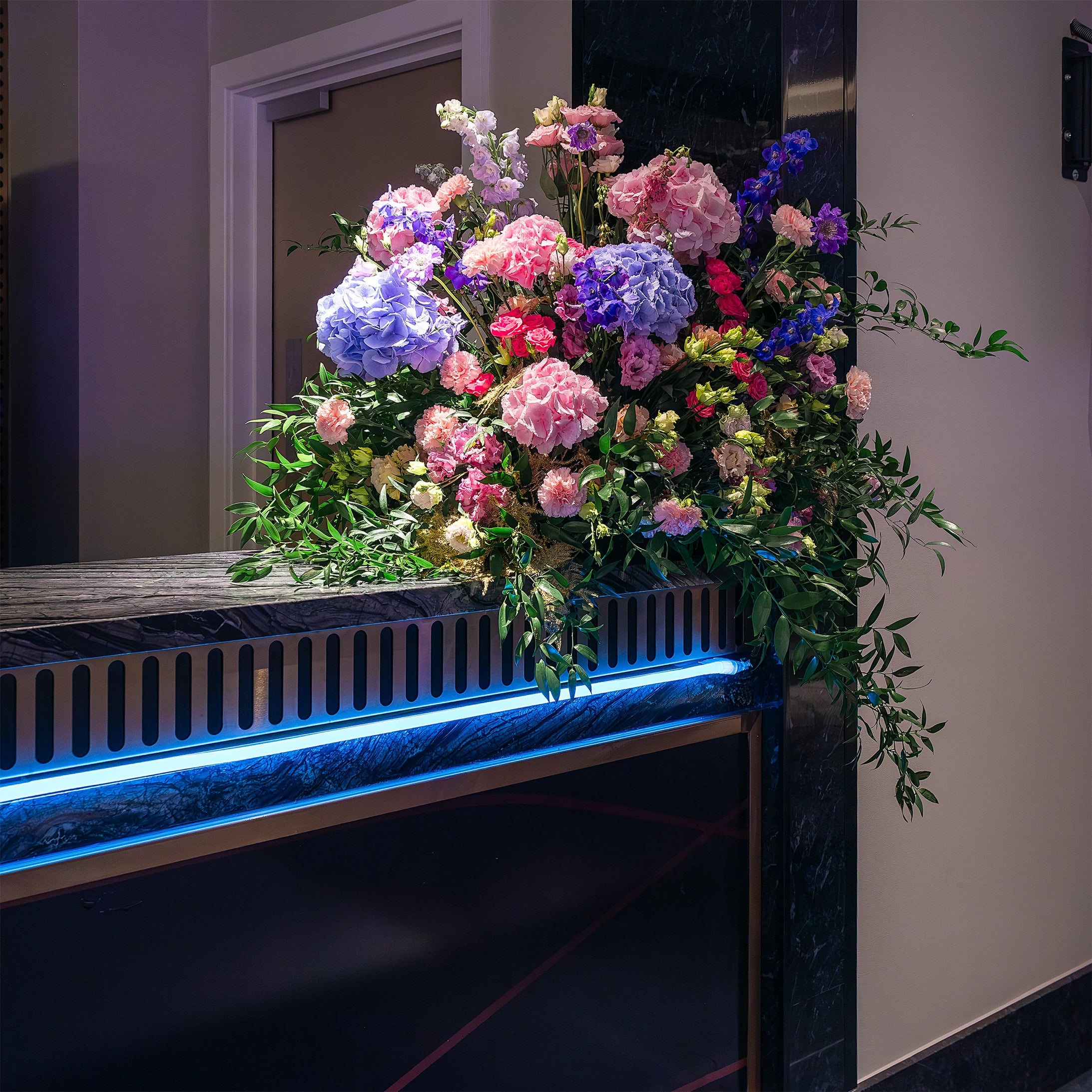 Exquisite floral arrangement on a reception counter, combining pastel roses, hydrangeas, and fresh greenery, highlighted by modern blue lighting, a signature of bespoke event floristry - Event Florist Amaranté London.