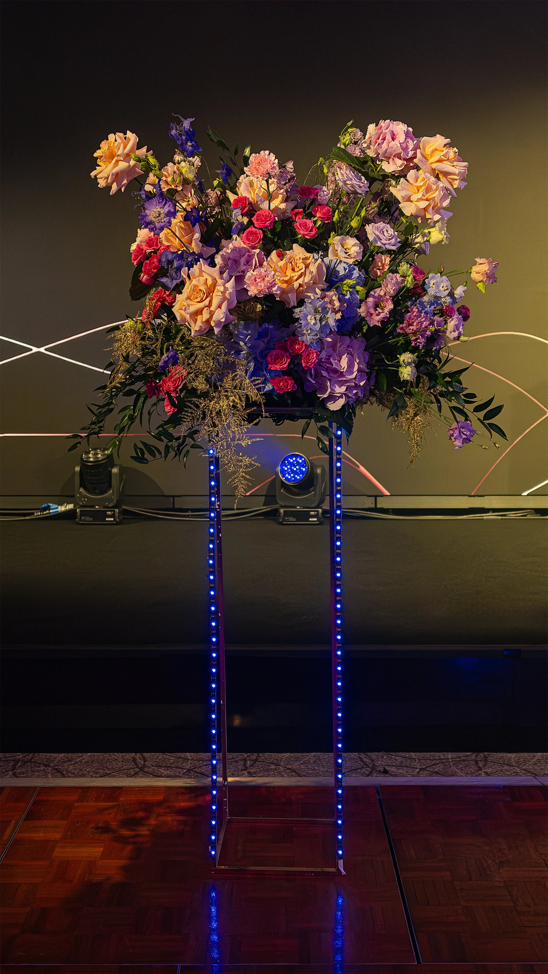 Tall floral arrangement on a sleek podium at the Formula E Awards Gala Dinner Event in London, featuring an opulent mix of pink roses, purple hydrangeas, and deep blue accents, highlighted by vertical neon blue lights - Amaranté London.