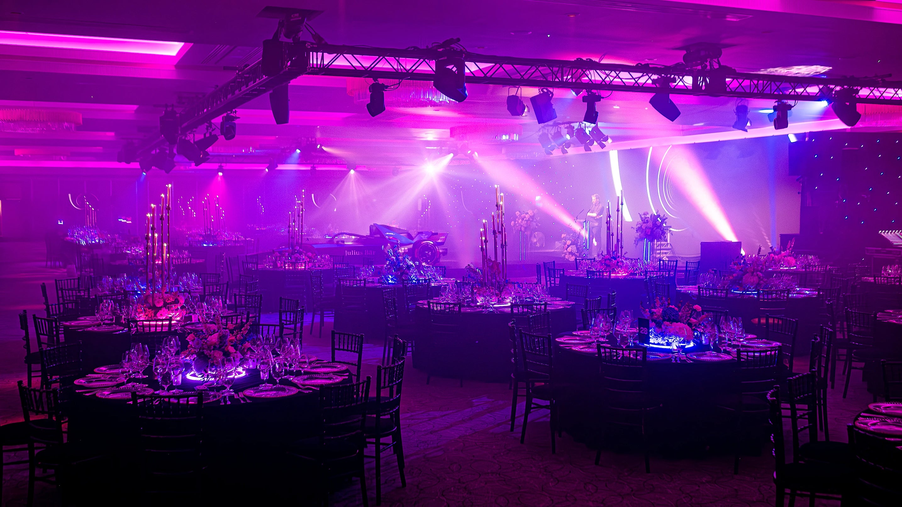 Expansive view of the Formula E Gala dinner setup with multiple tables adorned with rich floral arrangements, ambient purple lighting, and thematic neon accents - Amaranté London.