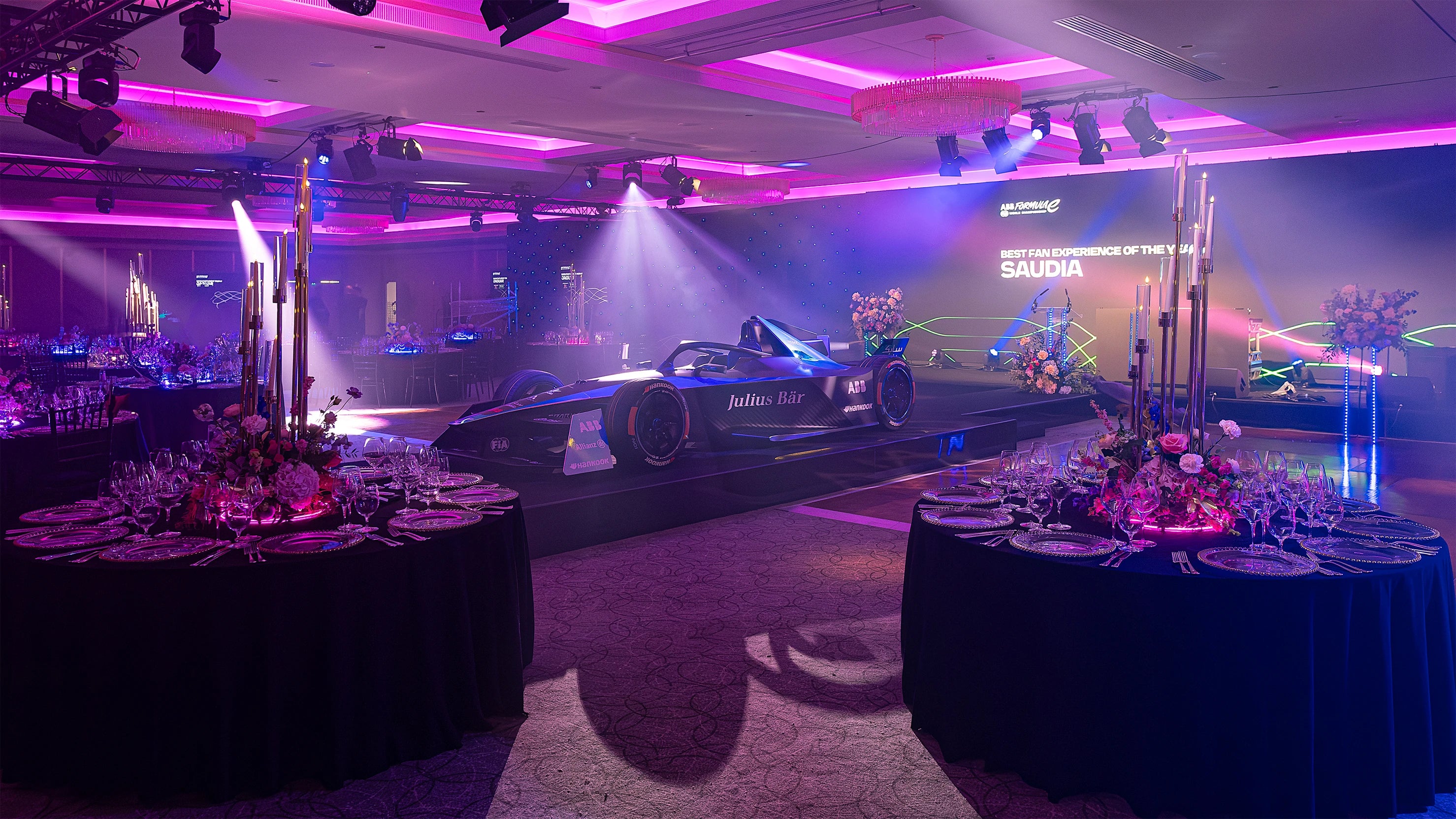 Formula E Gala dinner table with luxurious floral centrepieces, including roses, hydrangeas, and glowing tea lights, set against a backdrop of racecar displays and soft ambient lighting. Floral Design and flower arrangement creation by event florist Amaranté London.