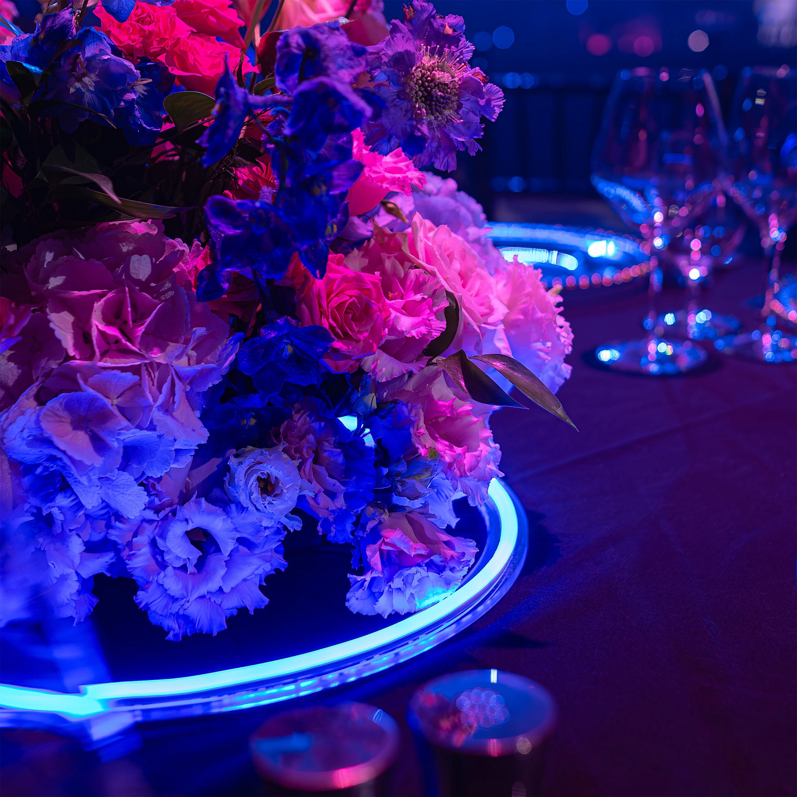An intimate table setting at the Formula E Gala featured dense, rich floral arrangements of blue and pink flowers, sophisticated glassware, and neon-lit decor, reflecting a high-end corporate event aesthetic - Amaranté London