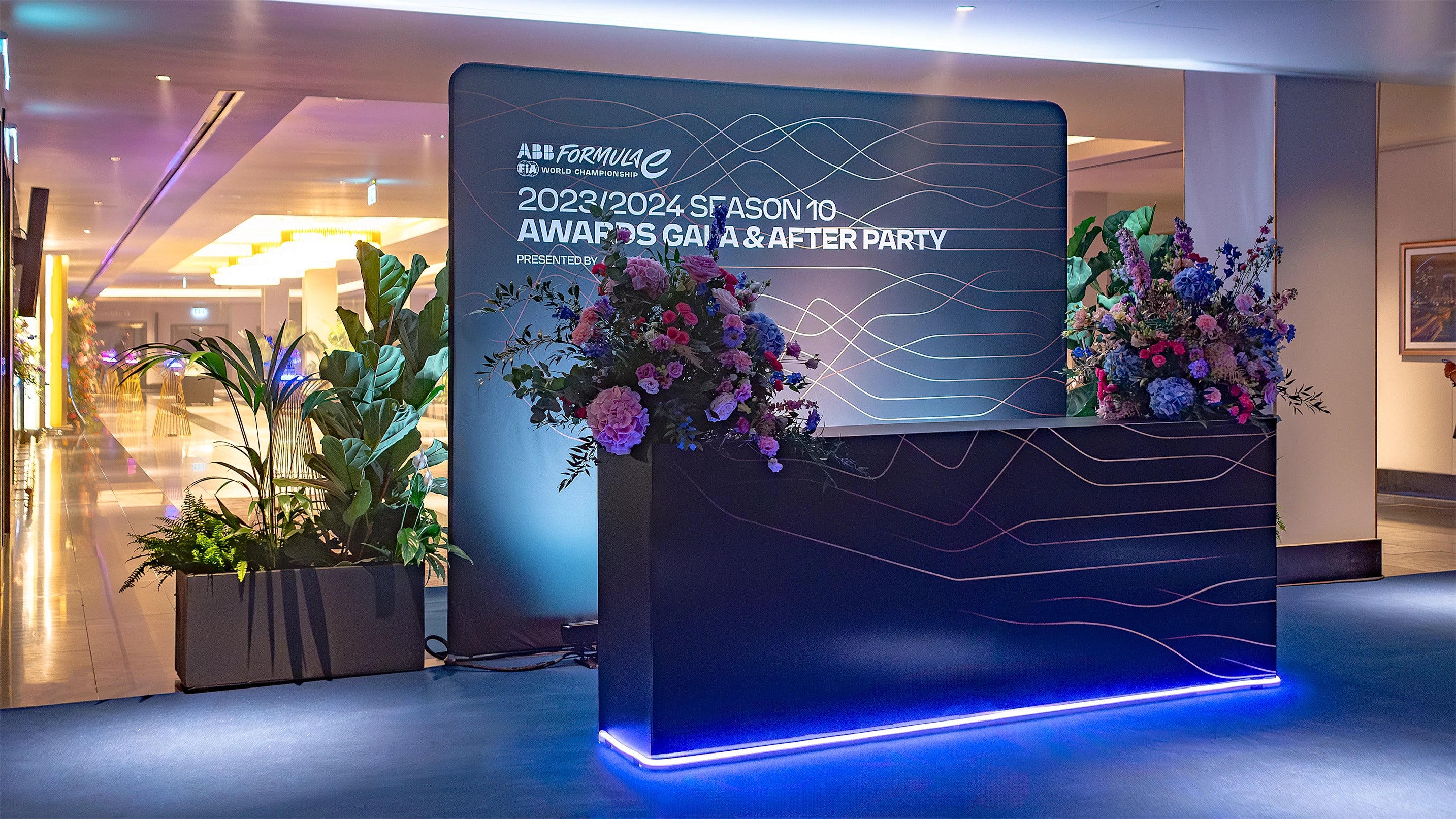 The stylish welcome area at the Formula E Gala Dinner in London is adorned with vibrant floral arrangements displayed on a sleek branded podium, surrounded by lush green plants, curated by Event Florist Amaranté London to create a memorable entrance for this event.