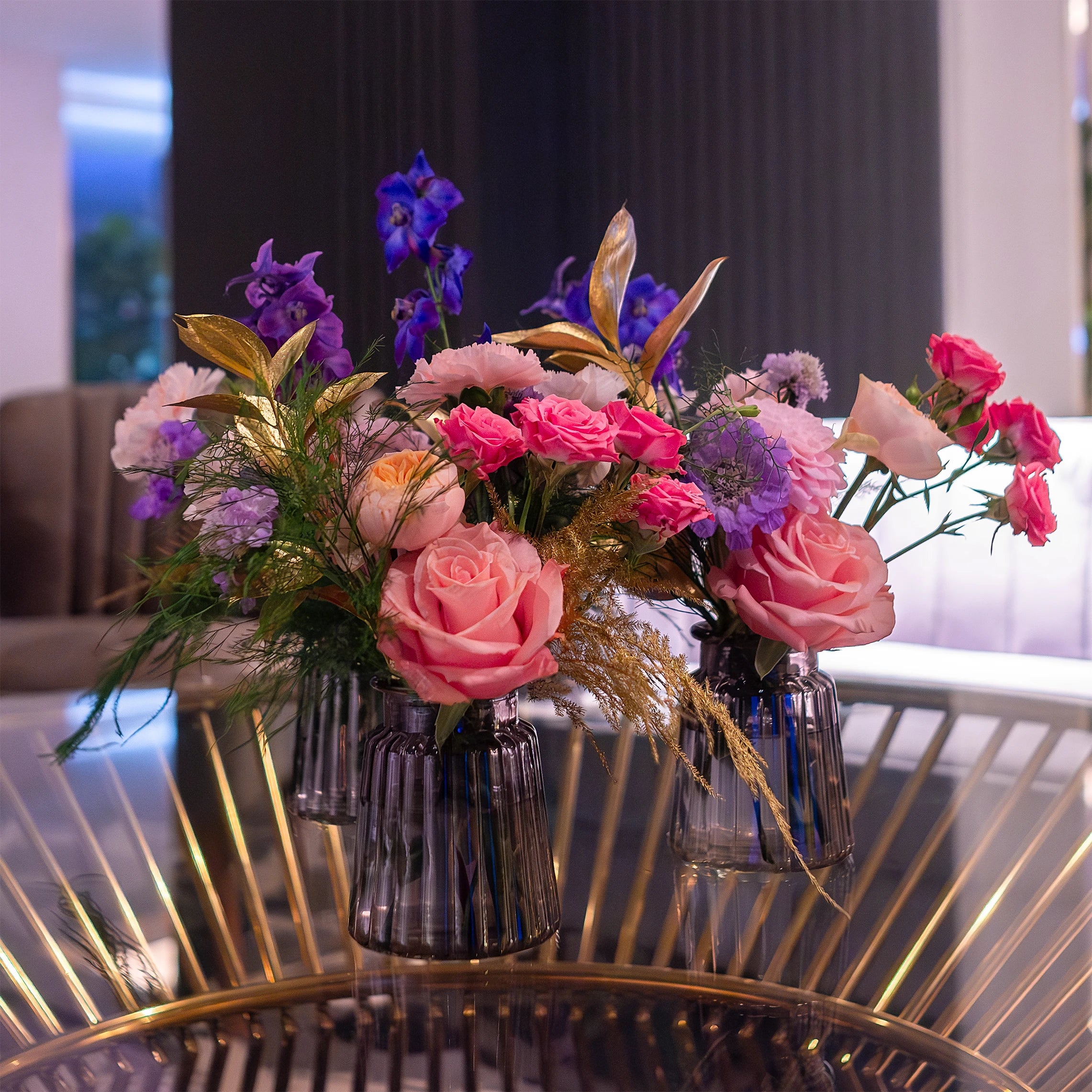 Chic floral centrepiece featuring roses in shades of pink and peach, blue irises, and golden accents in elegant glass vases on a gold metal table - Bespoke floral arrangements for events - Amaranté London, luxury event styling.