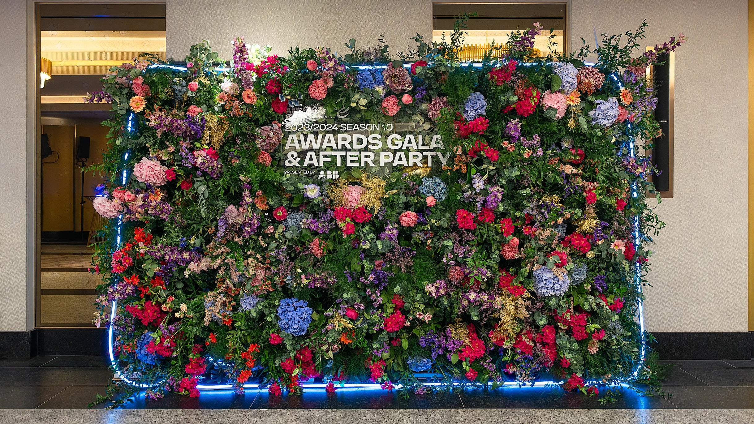 The expansive, bespoke luxury floral wall at the Formula E Awards Gala Dinner in London, featuring a lush array of pink, red, purple, and blue flowers, beautifully lit with soft blue lights - designed, created and installed by Event Florist Amaranté London.