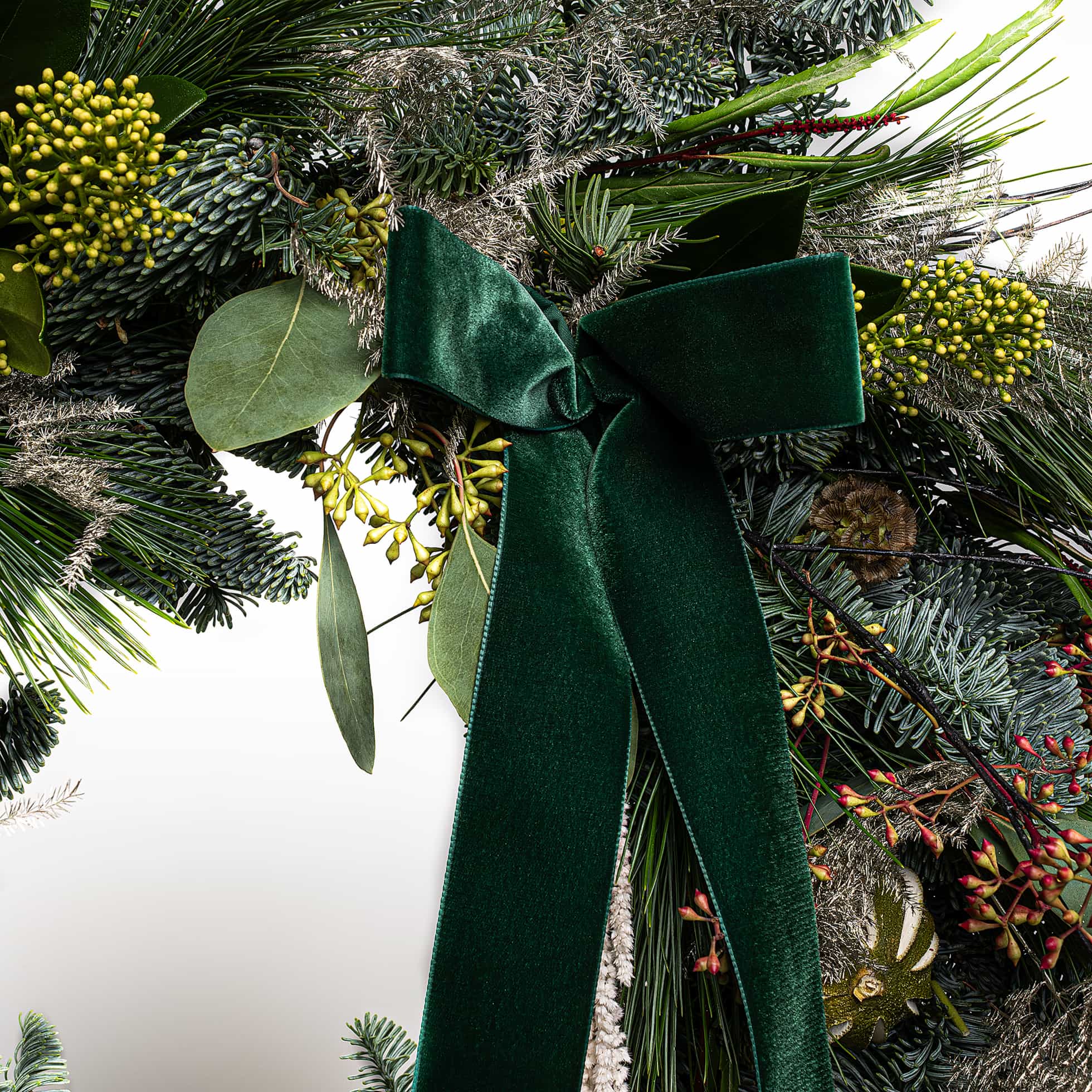 Close-up detail of luxury Christmas wreath featuring fresh dark green conifer sprigs, bright yellow-green flowering clusters, dark ornamental dried grasses, fresh eucalyptus leaves, white cascading dried strands, dark green velvet ribbon bow, and burgundy-toned dried botanical accents - Amaranté London