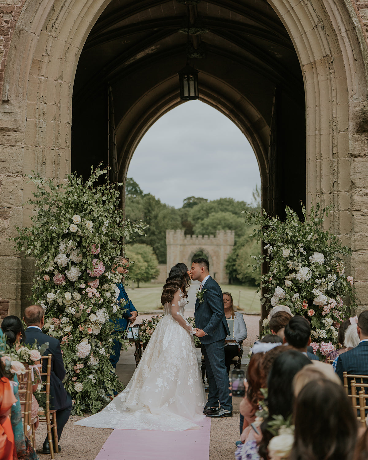 Wedding ceremony at Hampton Court Castle, Herefordshire, with bride and groom exchanging vows under a Gothic stone arch with floral arrangements and castle grounds visible - Floral Design by Amaranté London