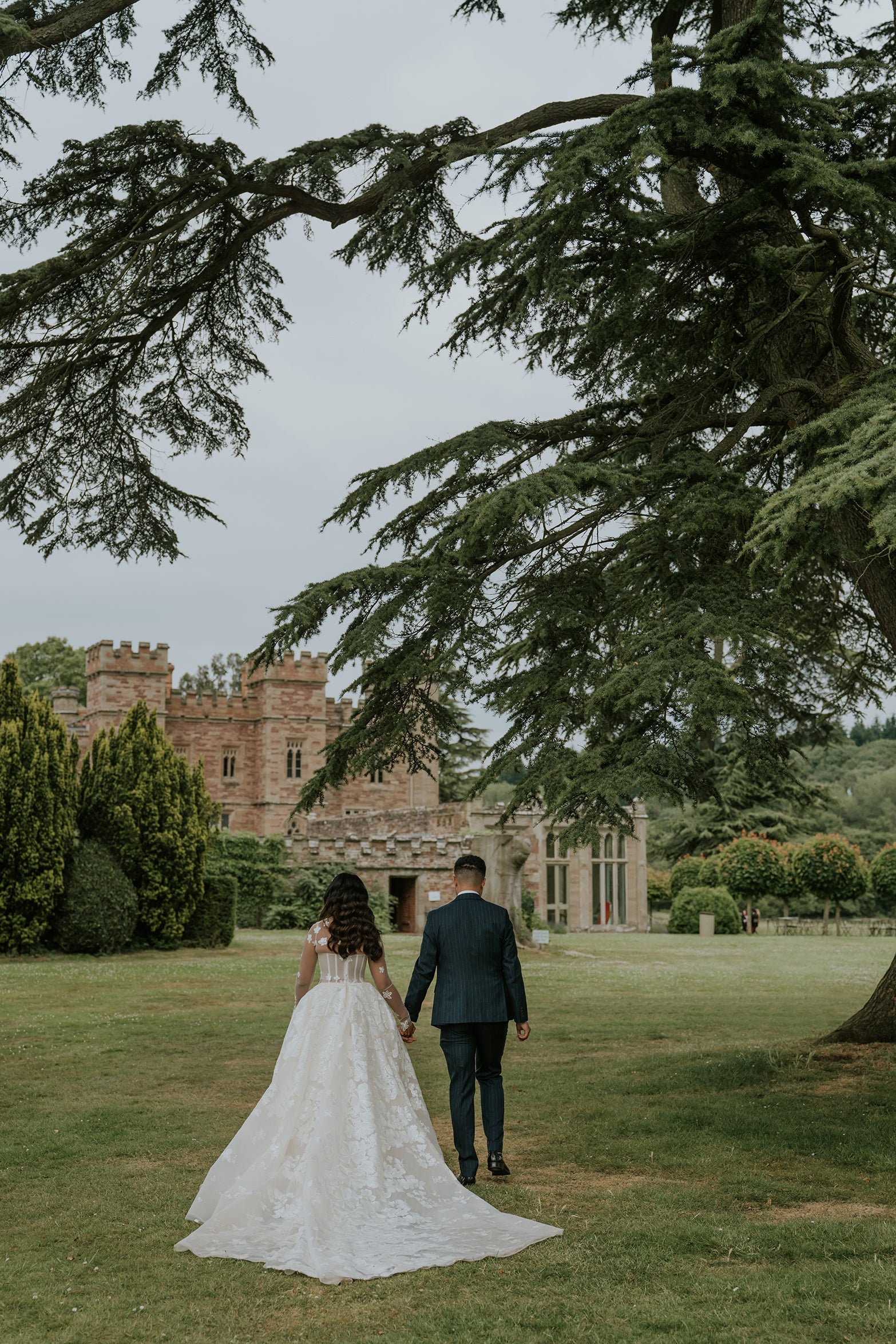 Bride and groom walking hand-in-hand across lawn toward Hampton Court Castle under large cedar tree - Floral Design by Amaranté London