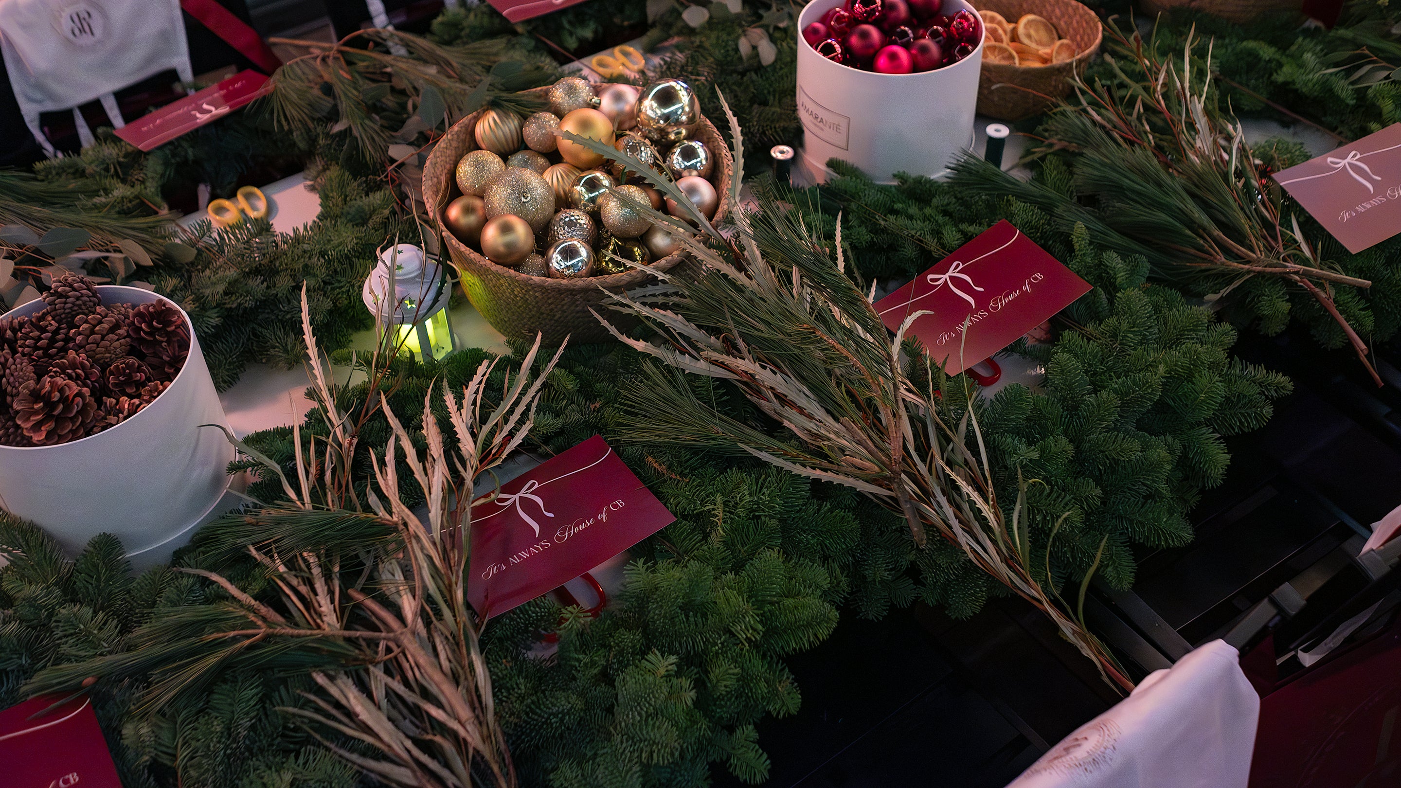 Overhead flatlay of luxury Christmas floristry styling featuring dried pampas grass and decorative foliage in jewel tones, gold and rose-gold baubles in a woven basket, burgundy Amaranté gift boxes, fresh evergreen sprigs, and white ceramic containers with festive ornaments and dried flowers arranged on a dark surface - Amaranté London